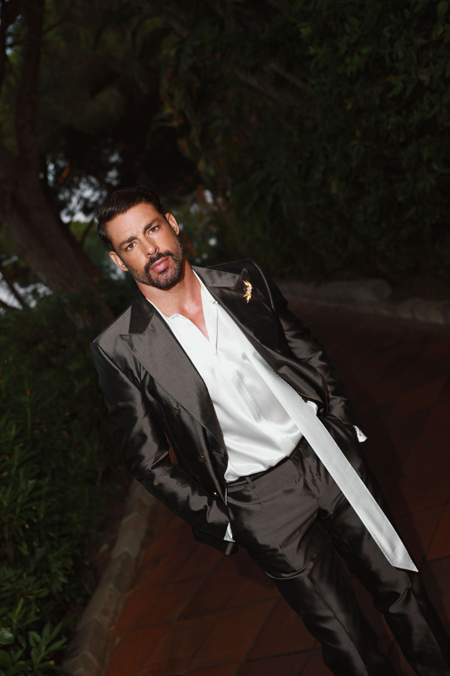 A person in a stylish dark suit and white shirt poses outdoors on a terracotta walkway near greenery.