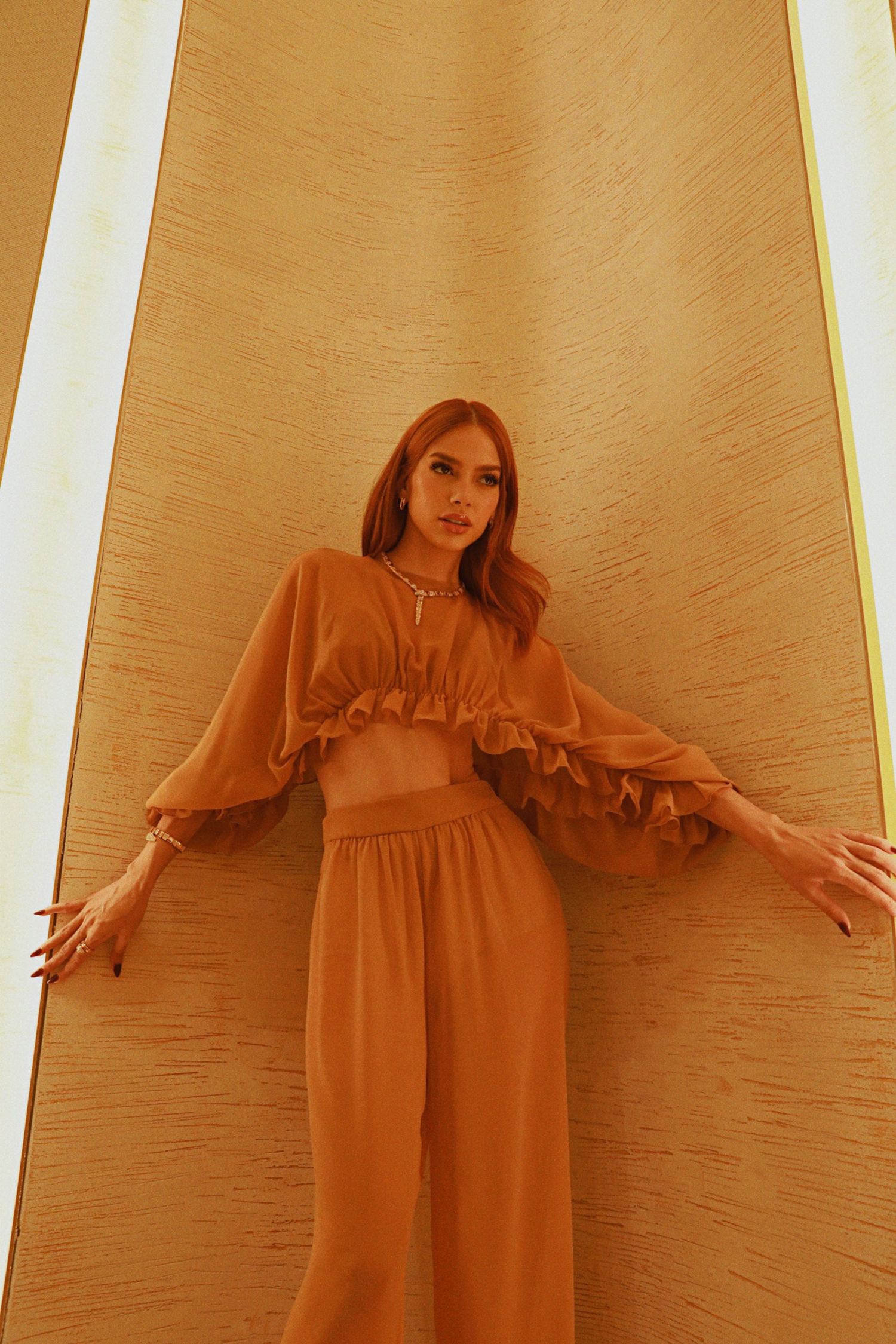Fashion model poses in flowing caramel-colored ensemble with ruffled crop top and wide-leg pants against textured wall.