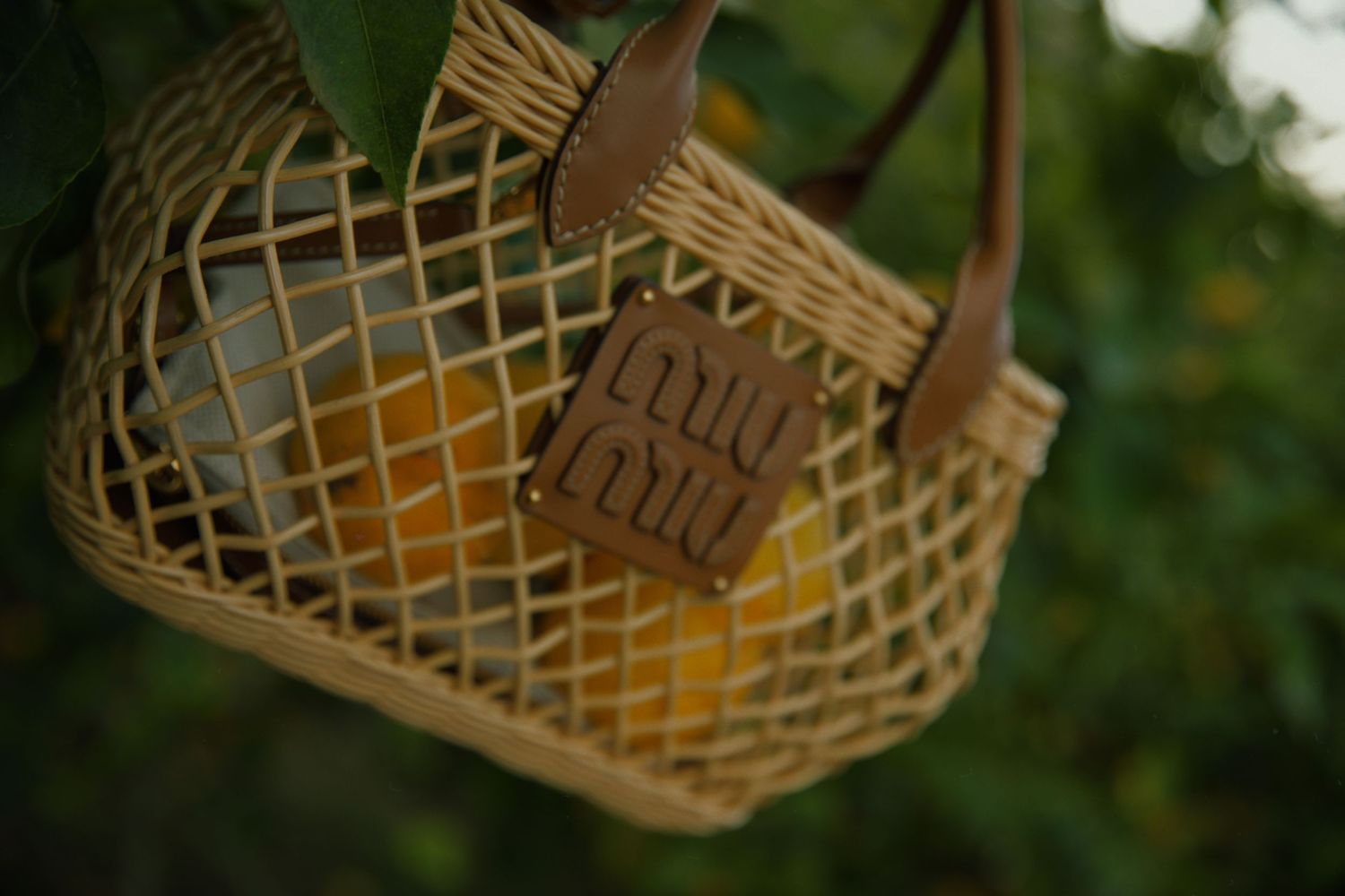 A close-up view of a woven bamboo basket with leather handles and tag hanging outdoors against greenery.