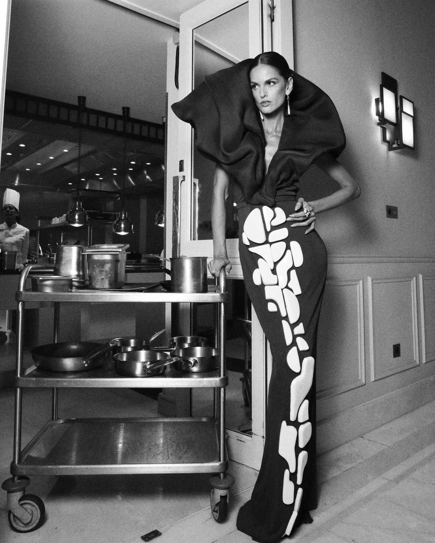 Dramatic black and white portrait of model in geometric dress beside industrial kitchen cart.