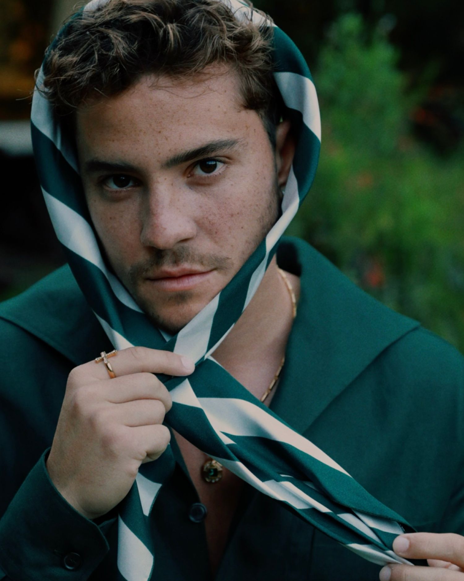 A person in a green hooded sweatshirt adjusts their striped scarf while posing for a casual outdoor portrait.