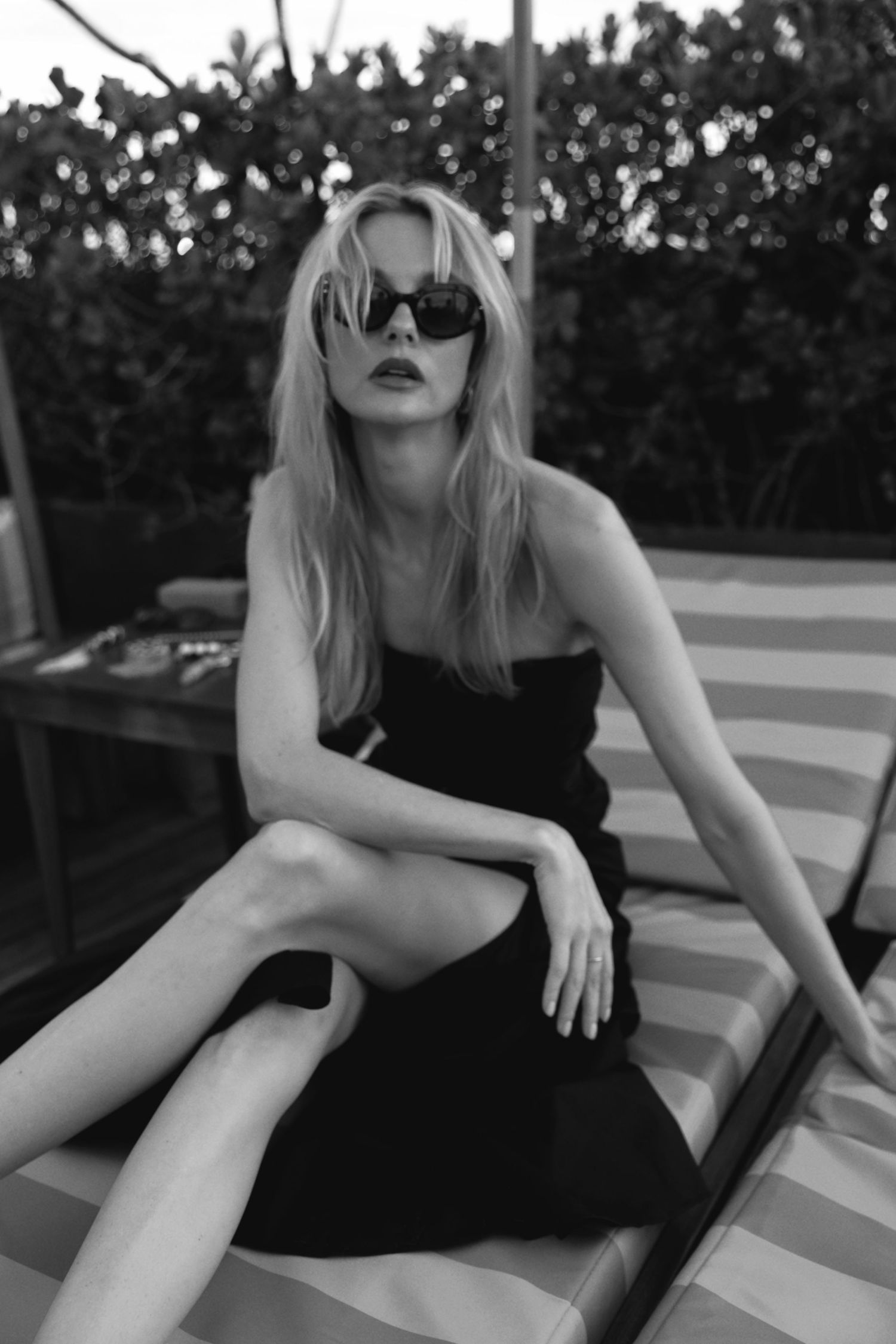 Black and white portrait of woman in sunglasses and black dress lounging outdoors.
