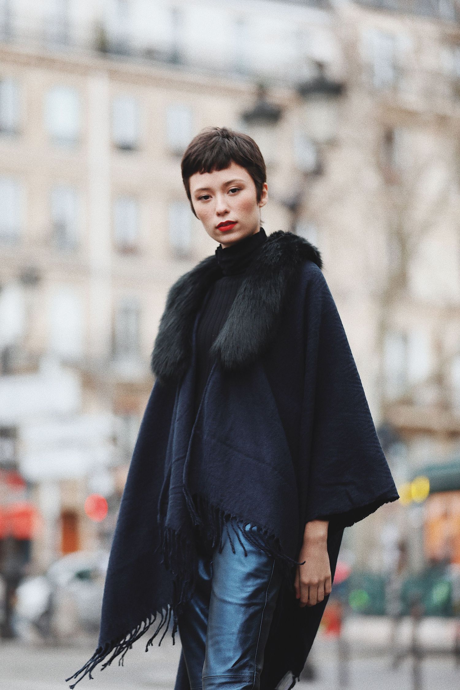 A fashionable street style look featuring a black poncho with fur trim and metallic blue pants on a city street.