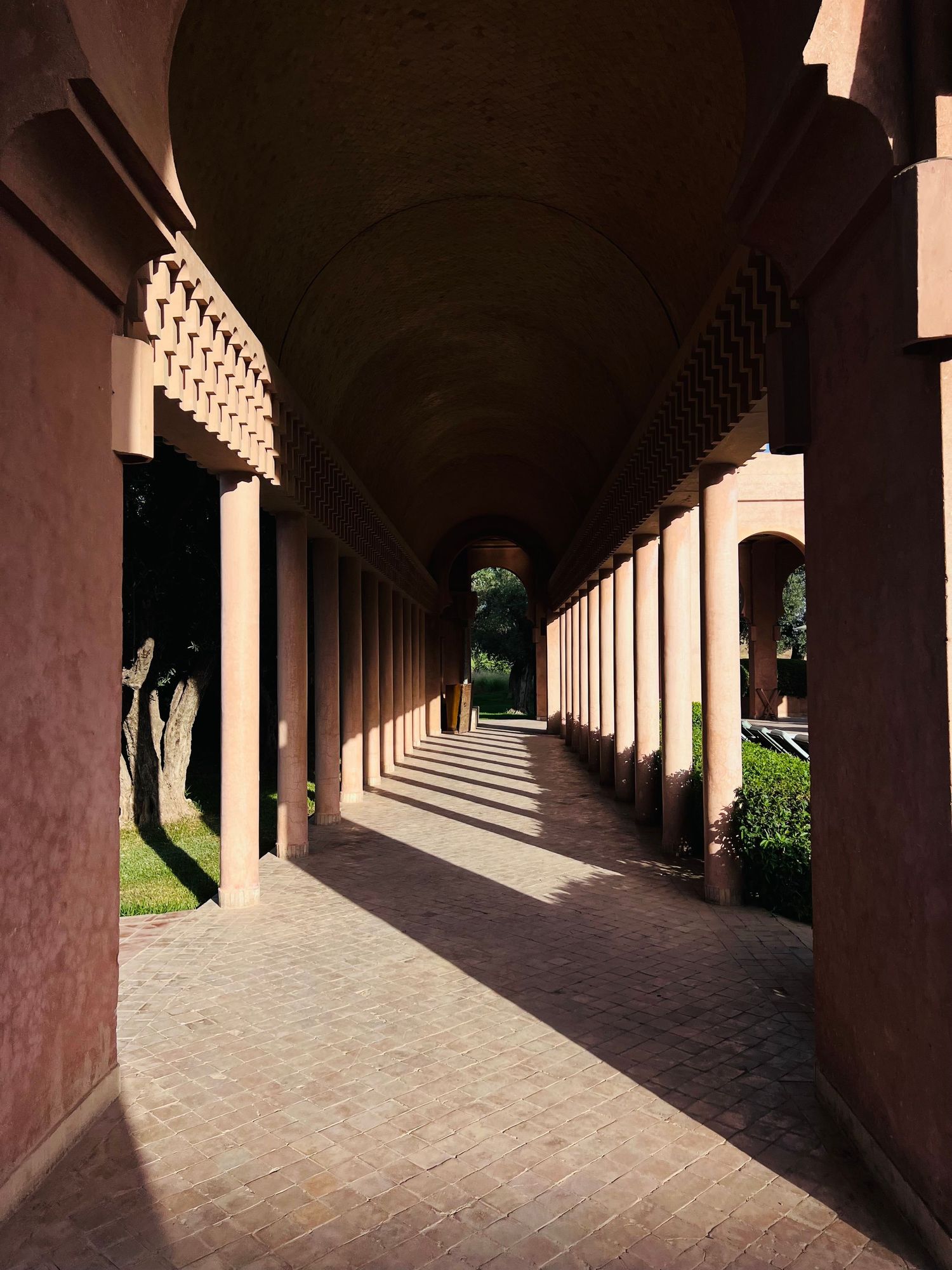 Sunlit arched colonnade walkway with terracotta pillars casting dramatic shadows along a brick paved path.