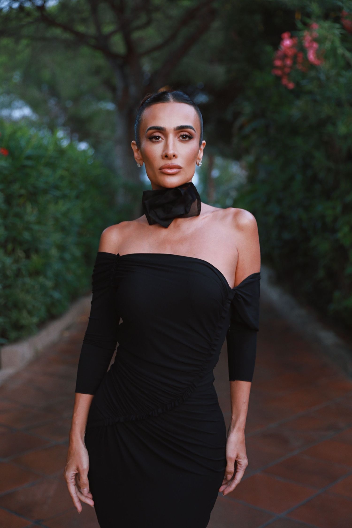 Model poses in black off-shoulder dress with choker necklace in garden setting.