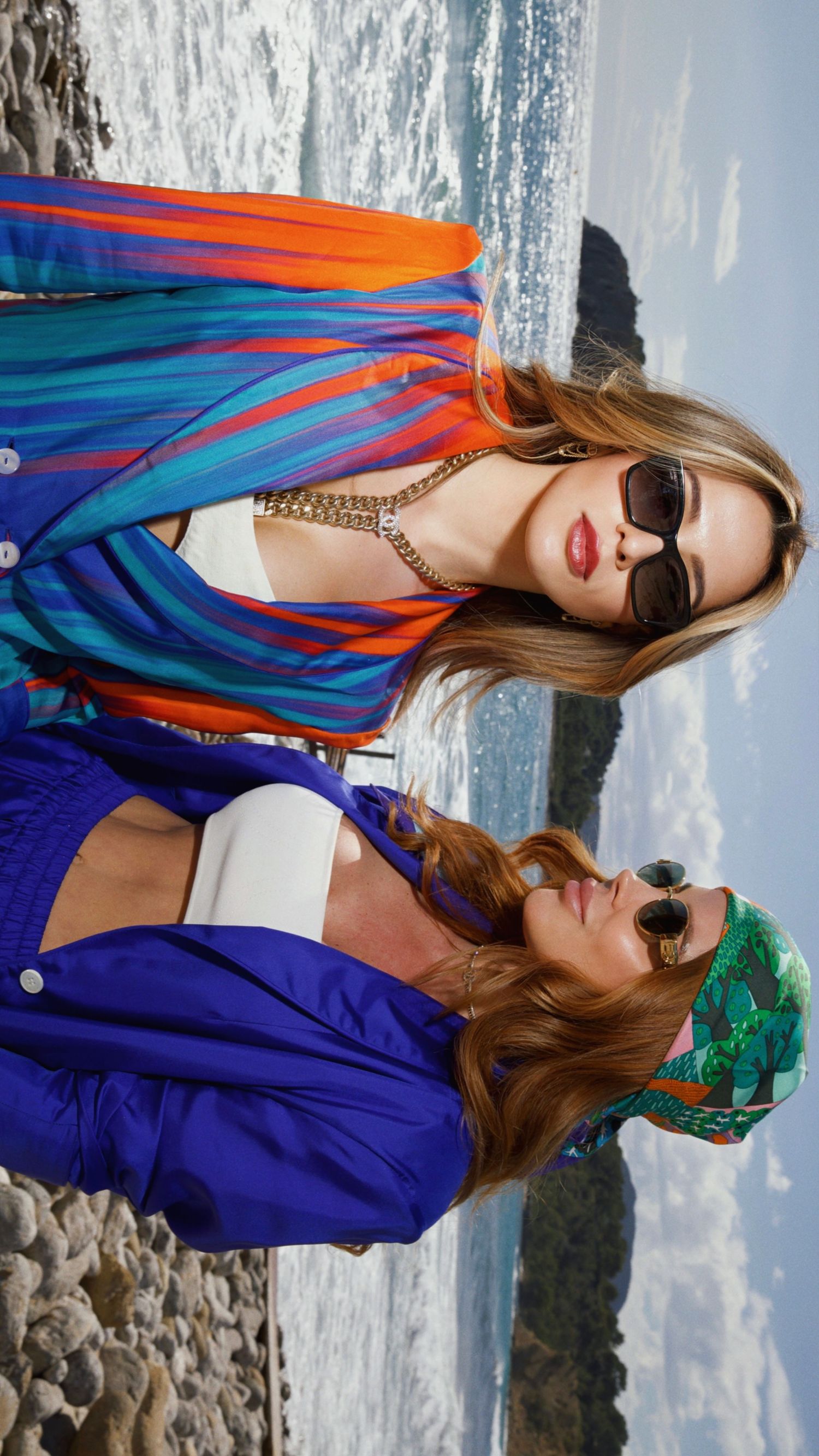 Two models in colorful resort wear posing by oceanside cliff in sunglasses.