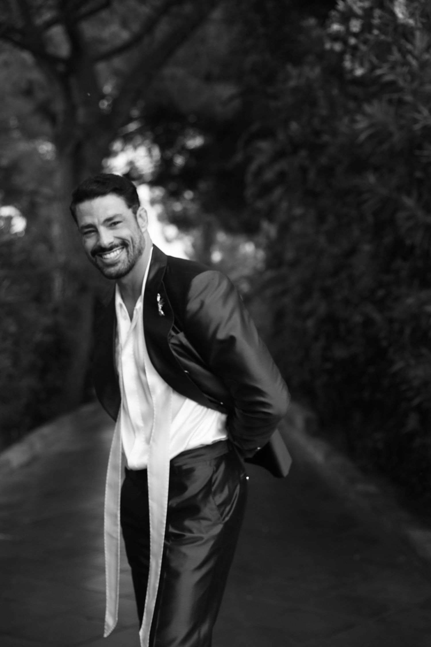 A black and white portrait of someone in a suit laughing against a blurred garden background.