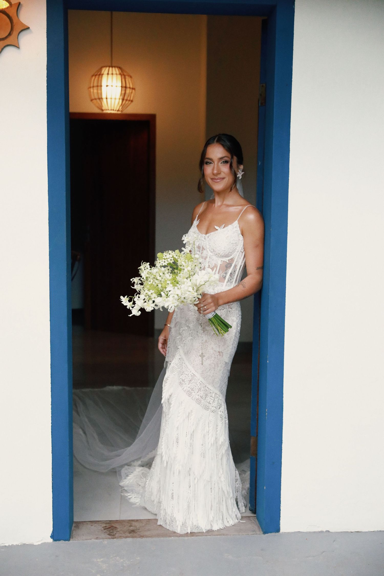 A bride in a fitted lace wedding gown stands in a blue doorframe holding a white floral bouquet.