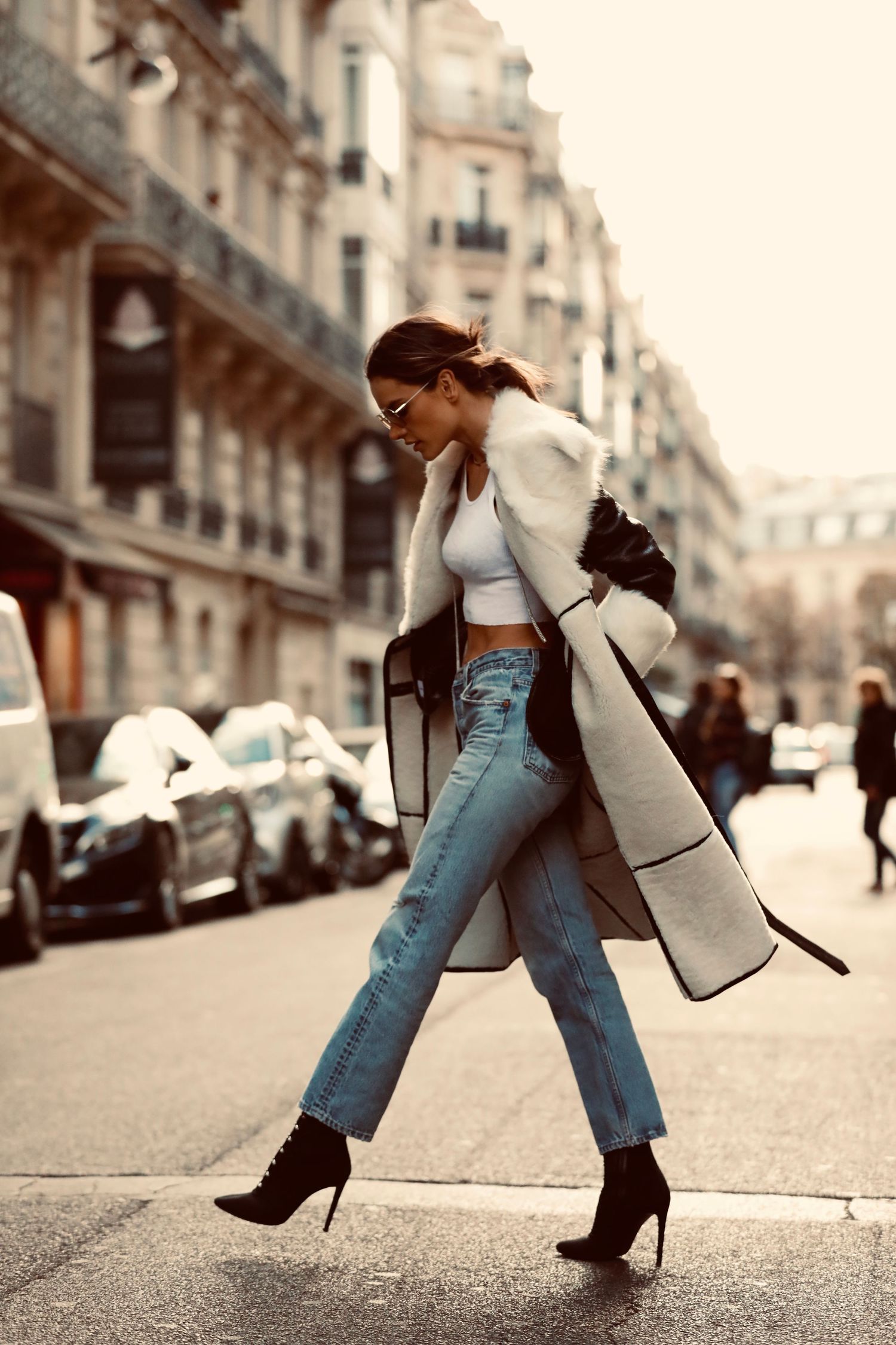 Stylish winter street style with long cream coat, cropped top, light blue jeans and black heels on a Paris street.