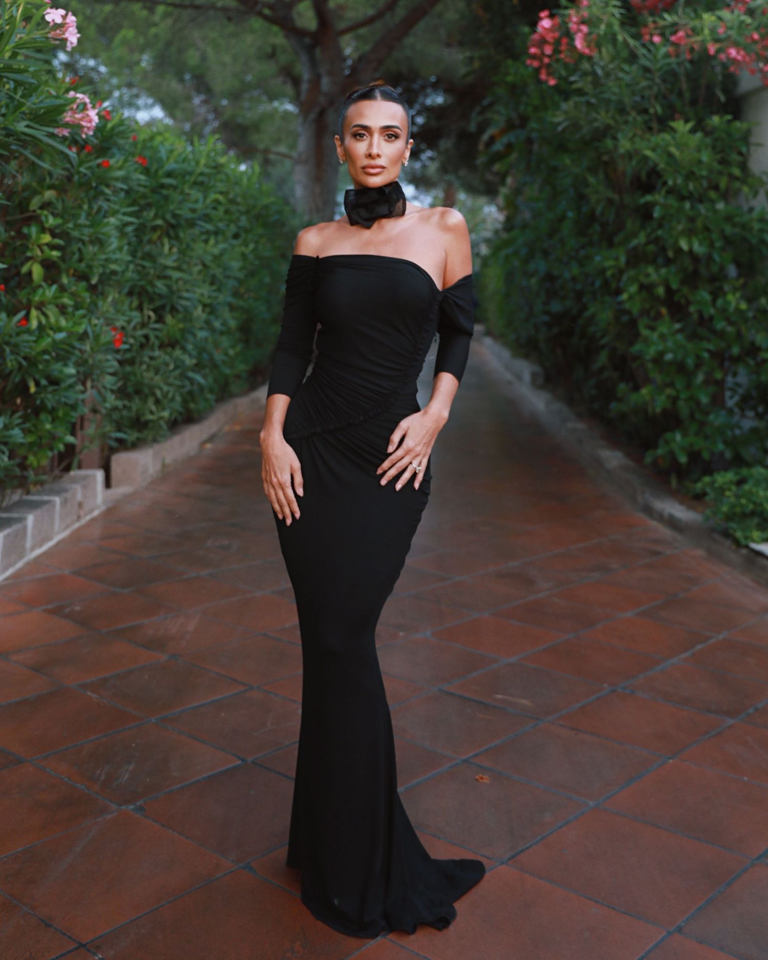 A model poses in an elegant black off-shoulder evening gown on a garden path with blooming flowers.