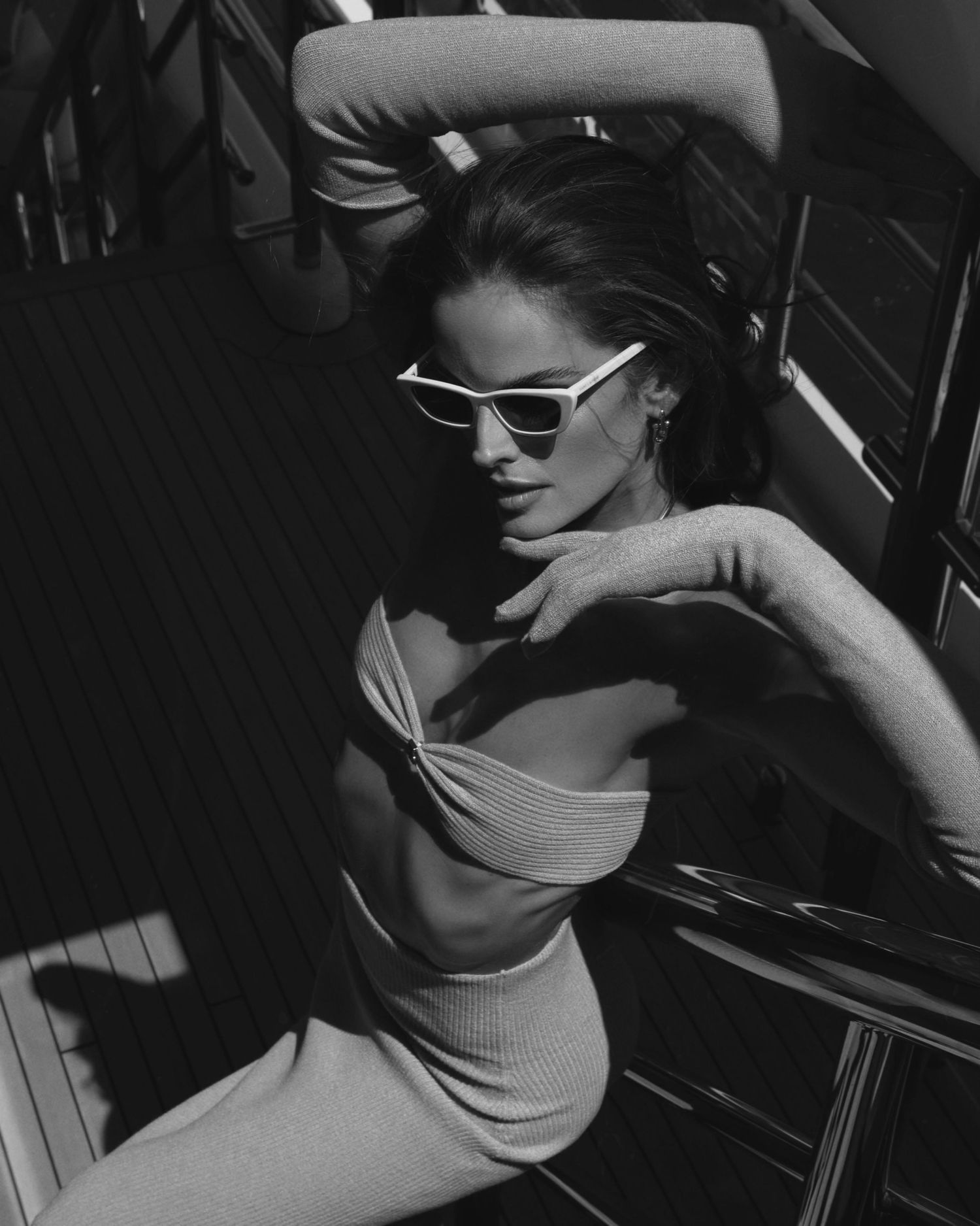 Artistic black and white portrait of woman in sunglasses and fitted outfit posing against dramatic shadows.