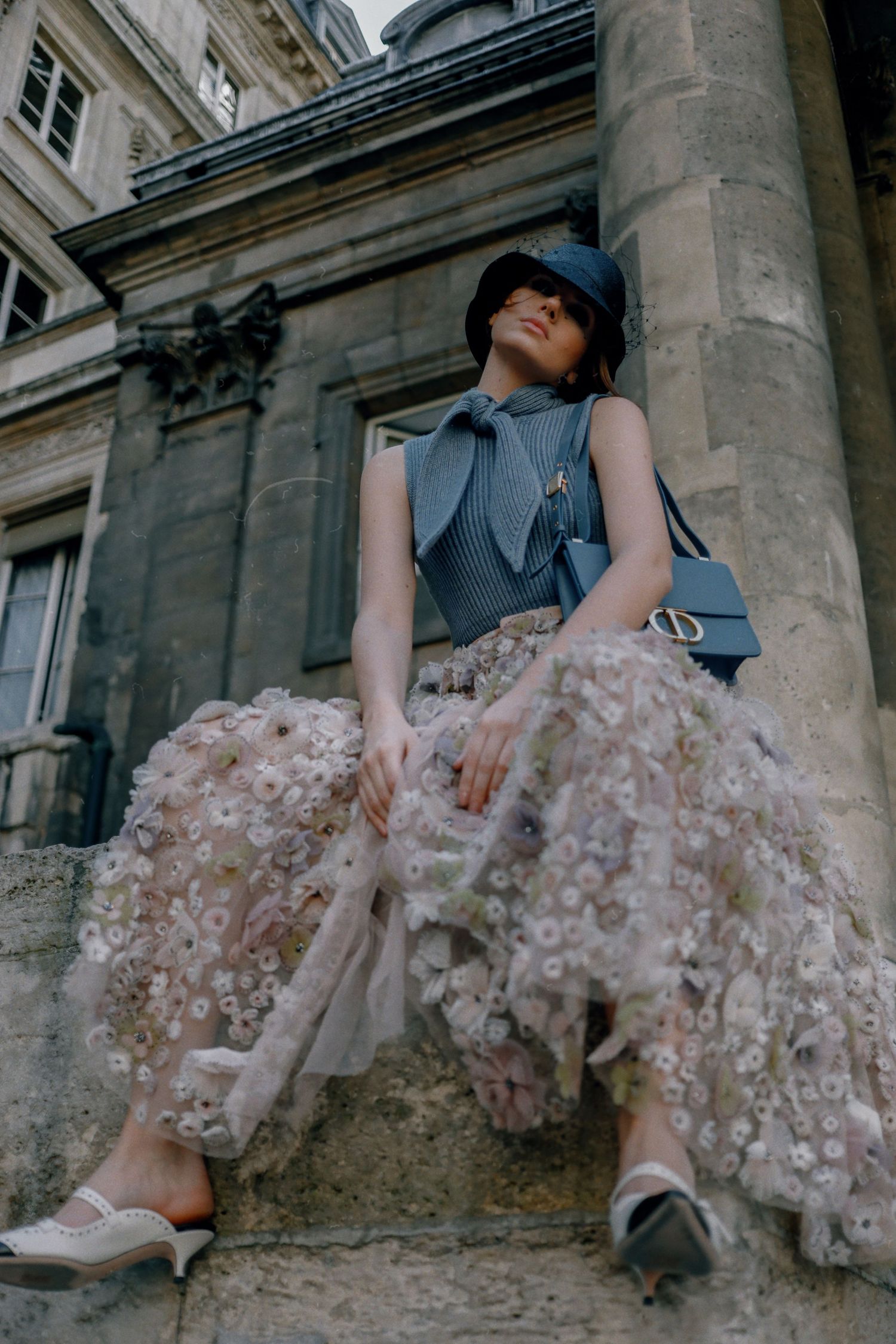 Fashion model in blue dress and hat sitting on stone steps wearing flowy tulle skirt with floral appliques.