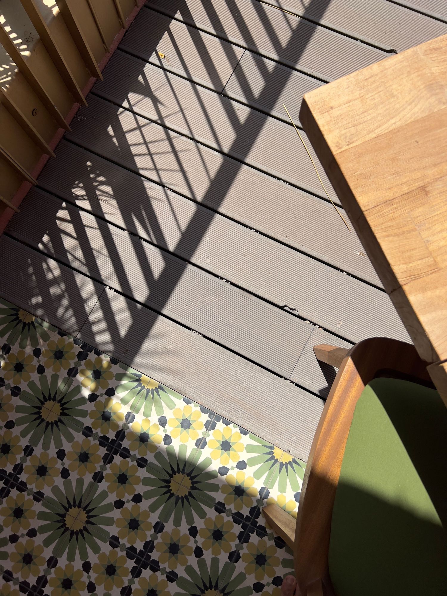 Sunlight casts dramatic diagonal shadows through window blinds onto gray carpet near a patterned floor and wooden furniture.