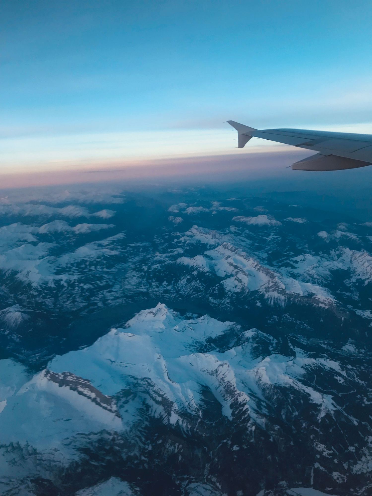 Aerial view of snow-capped mountains beneath airplane wing during sunset with vibrant blue and orange sky.