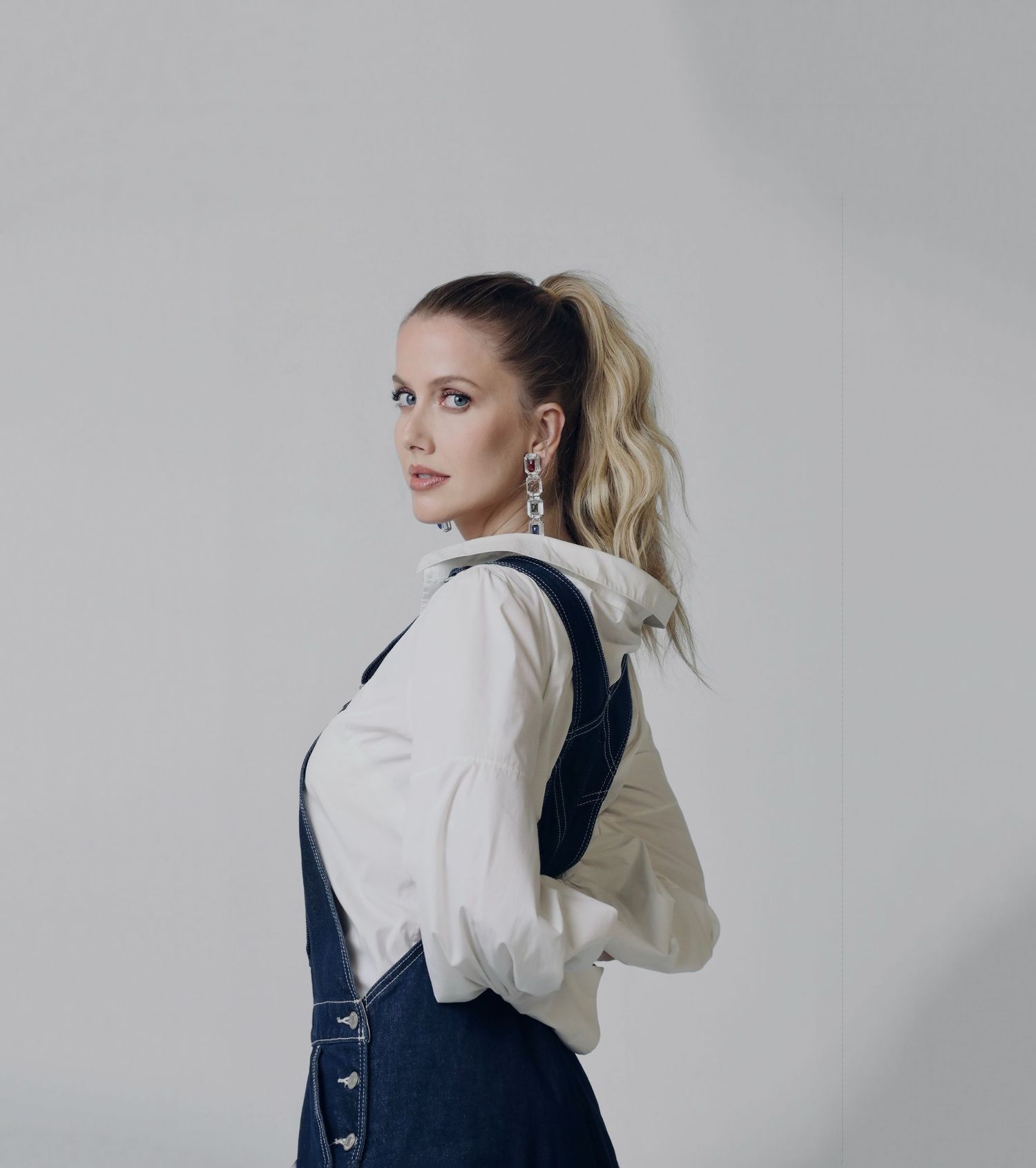 Side profile of model in white blouse and navy overalls against gray background.