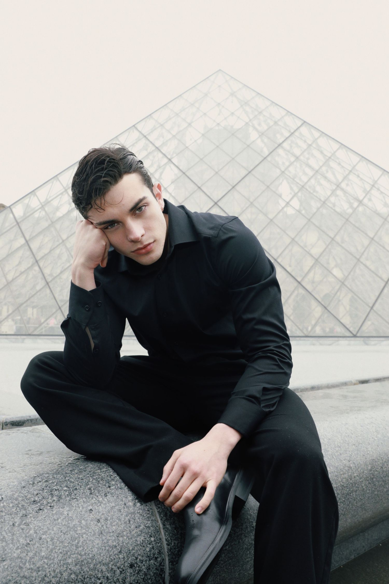 Model in all-black outfit poses thoughtfully near geometric glass pyramid structure.