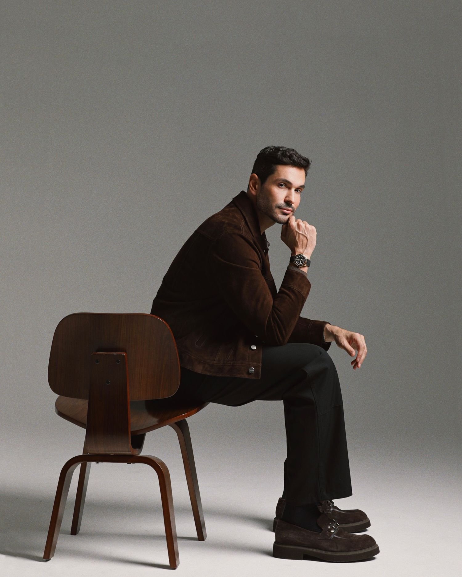 Figure in brown corduroy jacket seated on wooden chair against gray backdrop in minimalist fashion photoshoot.