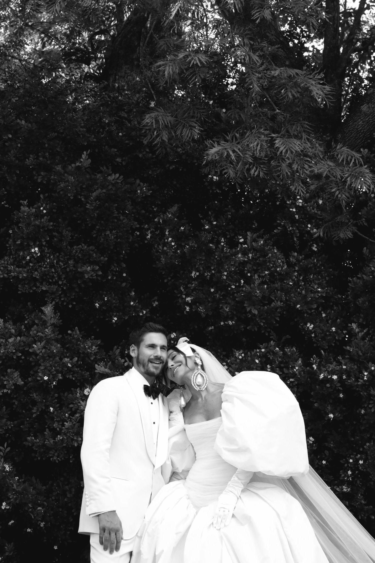 Black and white wedding photo showing couple in white attire embracing outdoors against lush greenery backdrop.