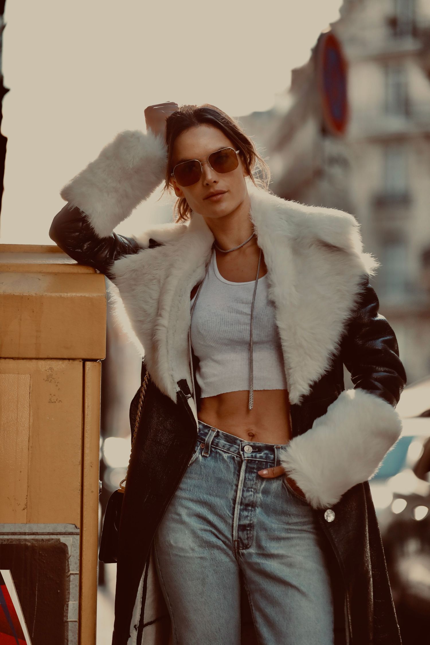 Person in stylish winter outfit with black leather jacket, white fur trim, crop top and high-waisted jeans poses outdoors.
