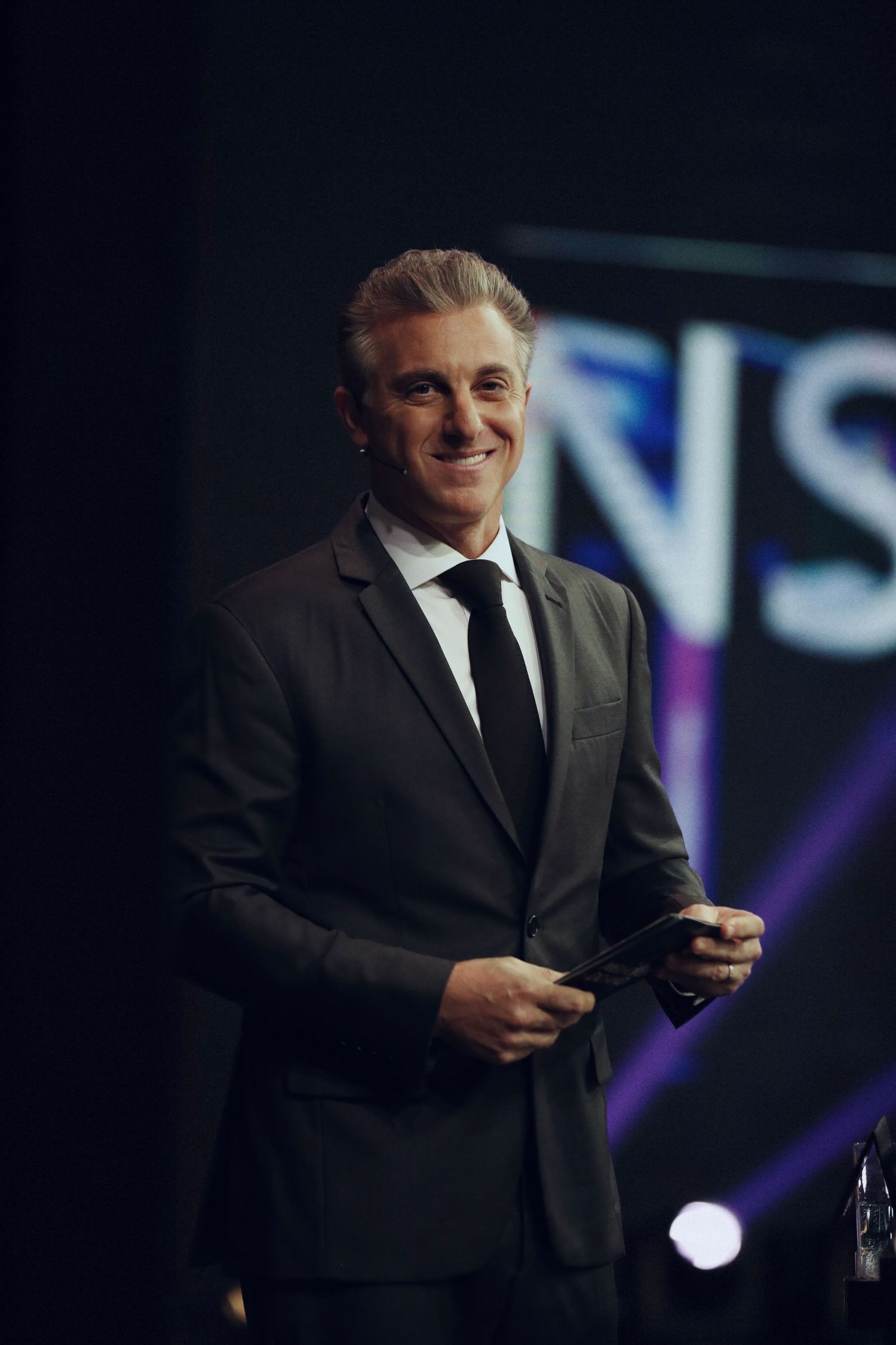 Professional shot of a person in formal suit speaking at an event with stage lighting.