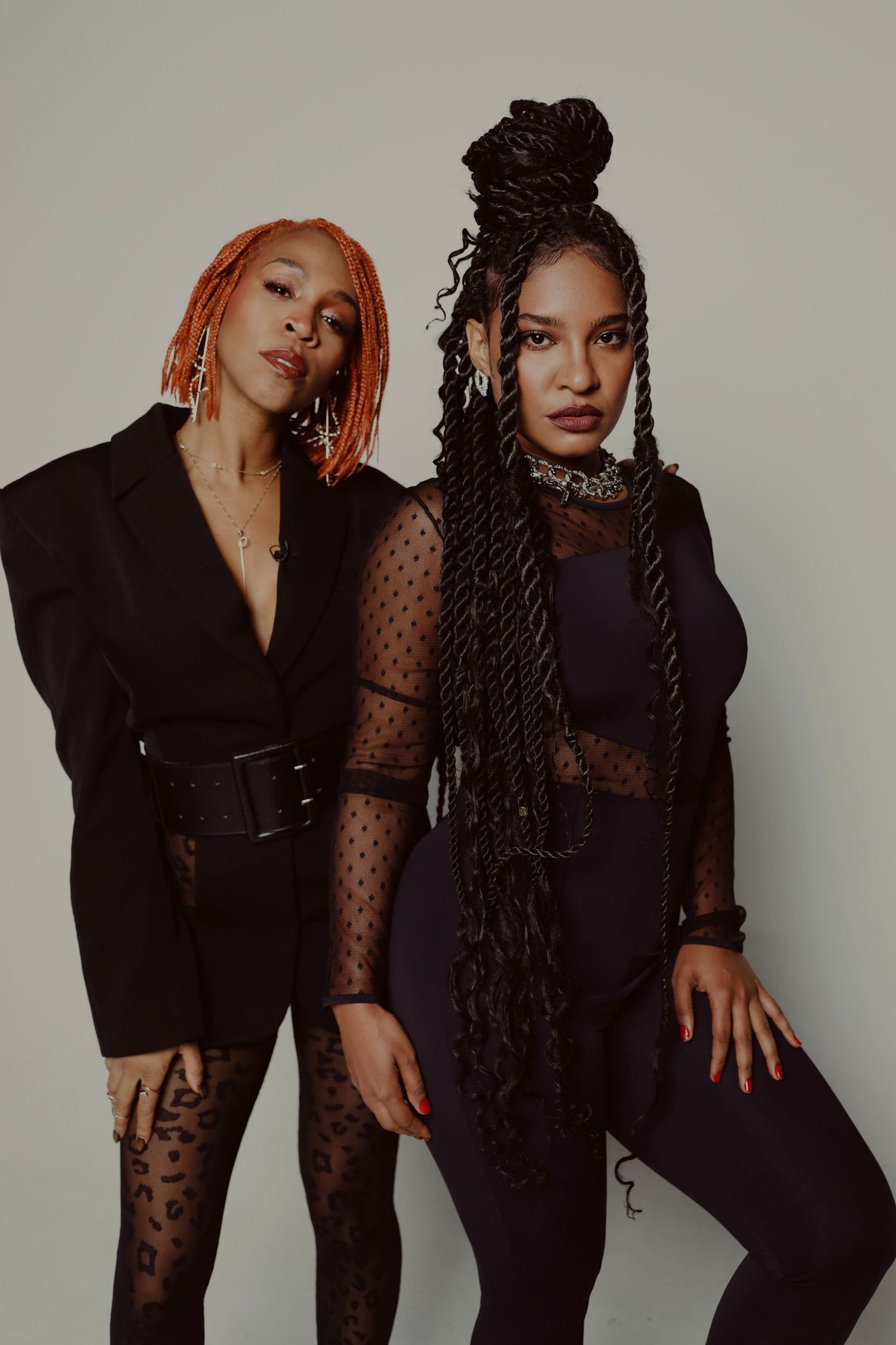 Two stylish figures pose in black outfits with unique hairstyles against neutral background.