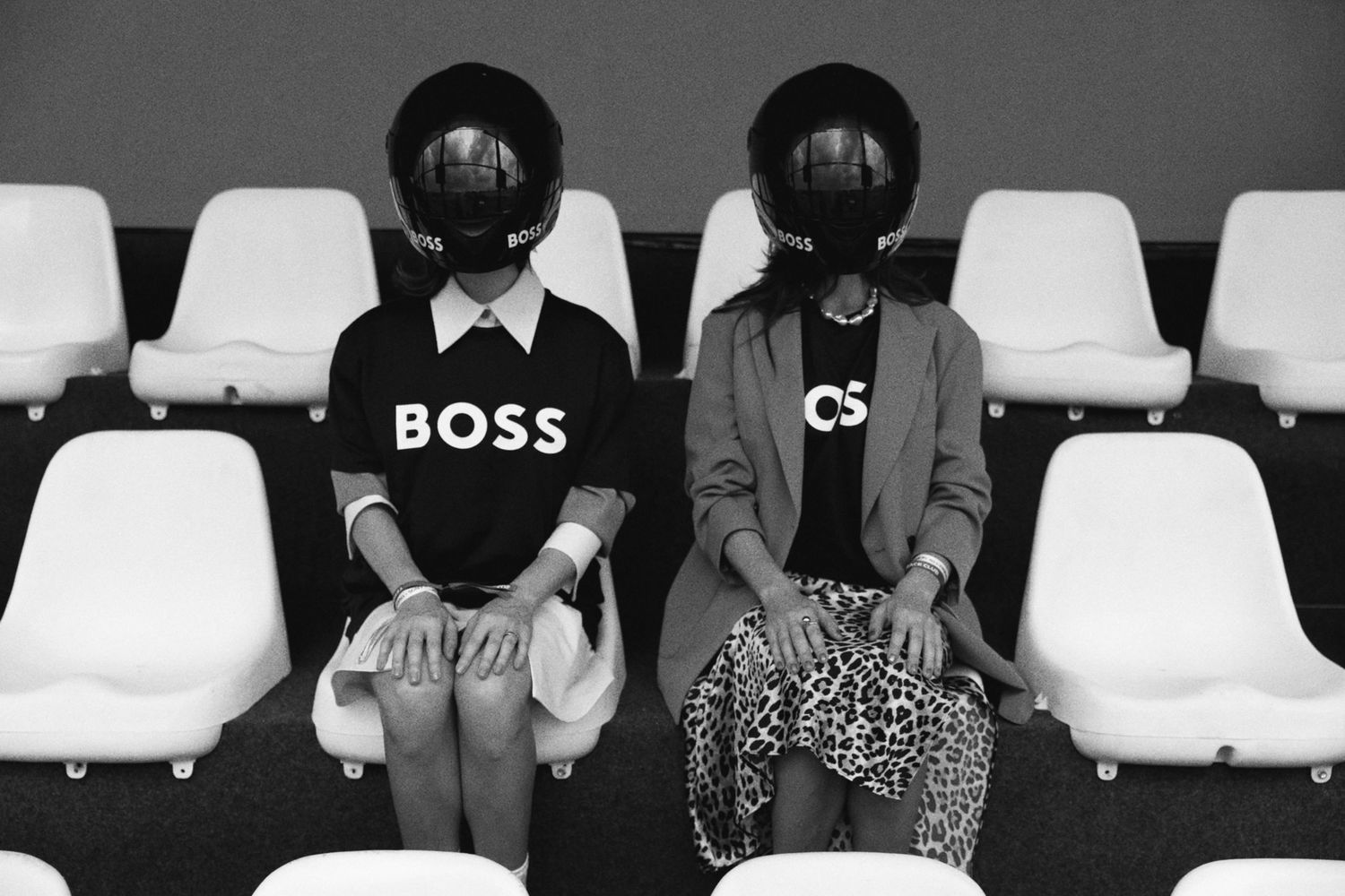 A black and white artistic photo of two figures in stadium seats wearing round helmets, one in a BOSS shirt.