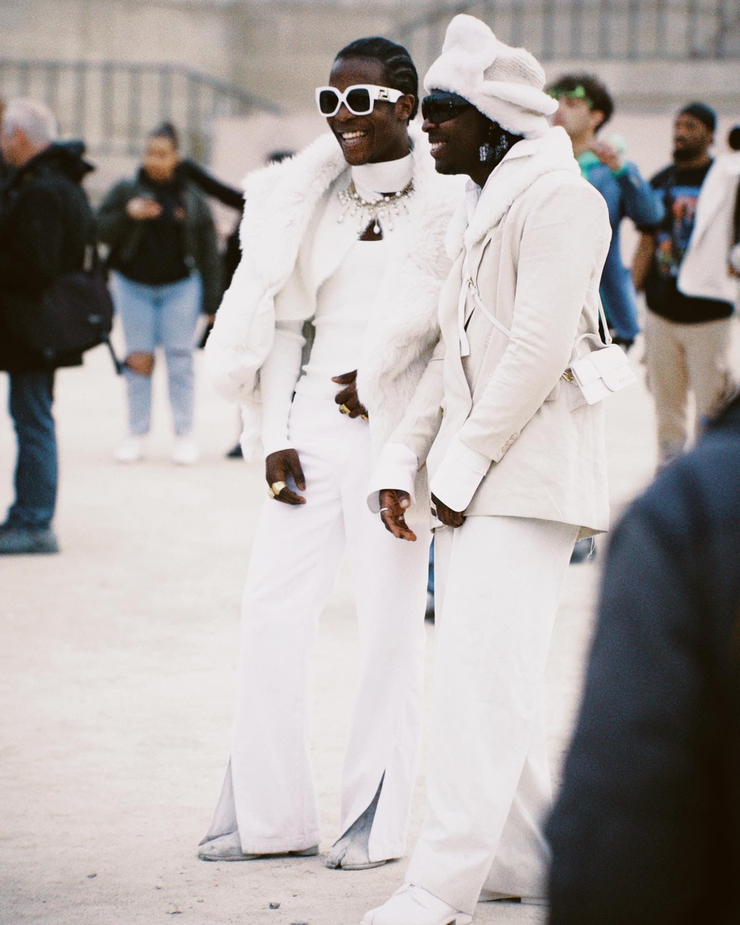 Two people in stylish all-white outfits and sunglasses stand together at an outdoor urban gathering.