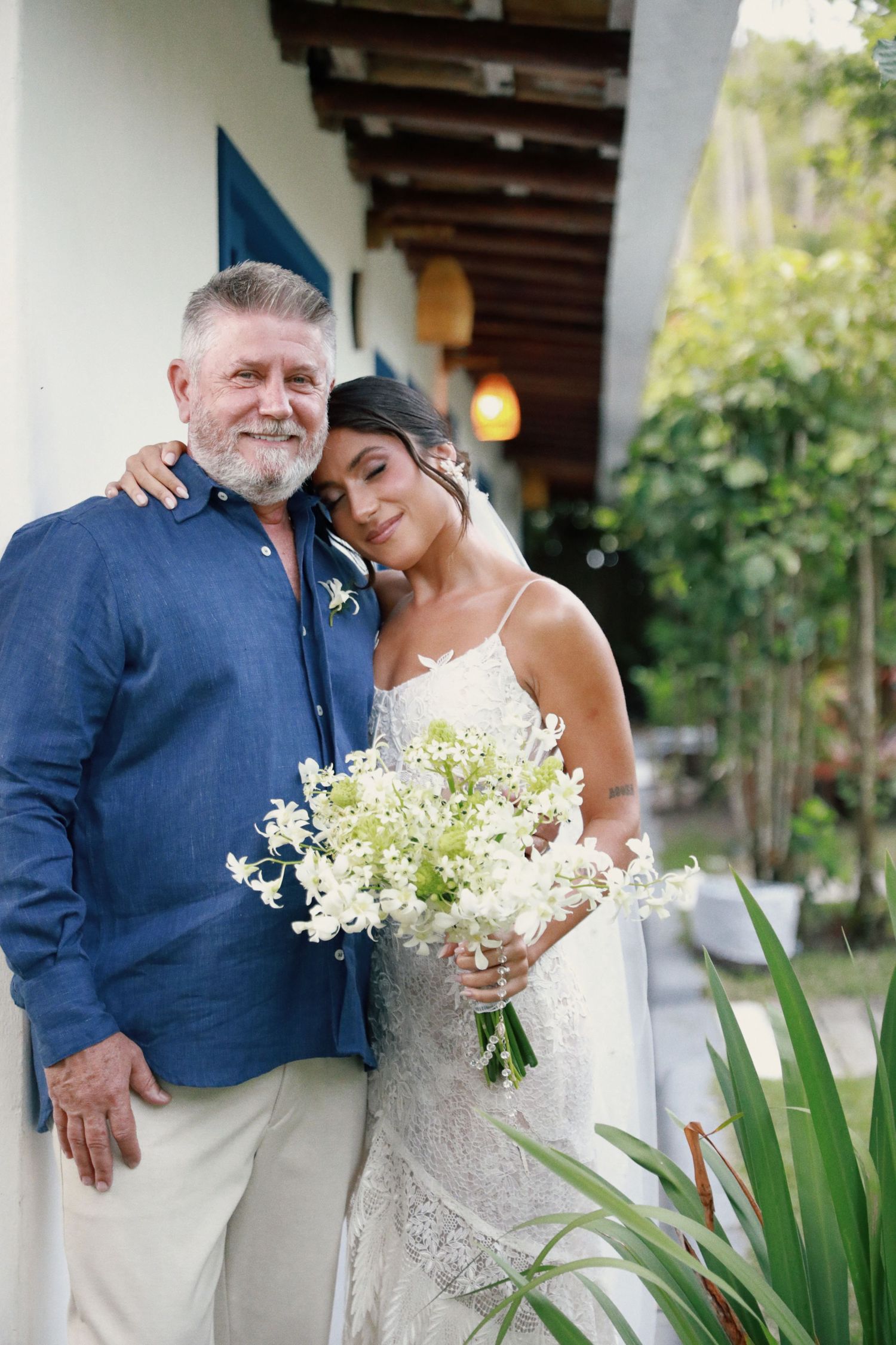A couple shares a tender moment at a tropical wedding venue, with the bride holding a delicate bouquet of white flowers.