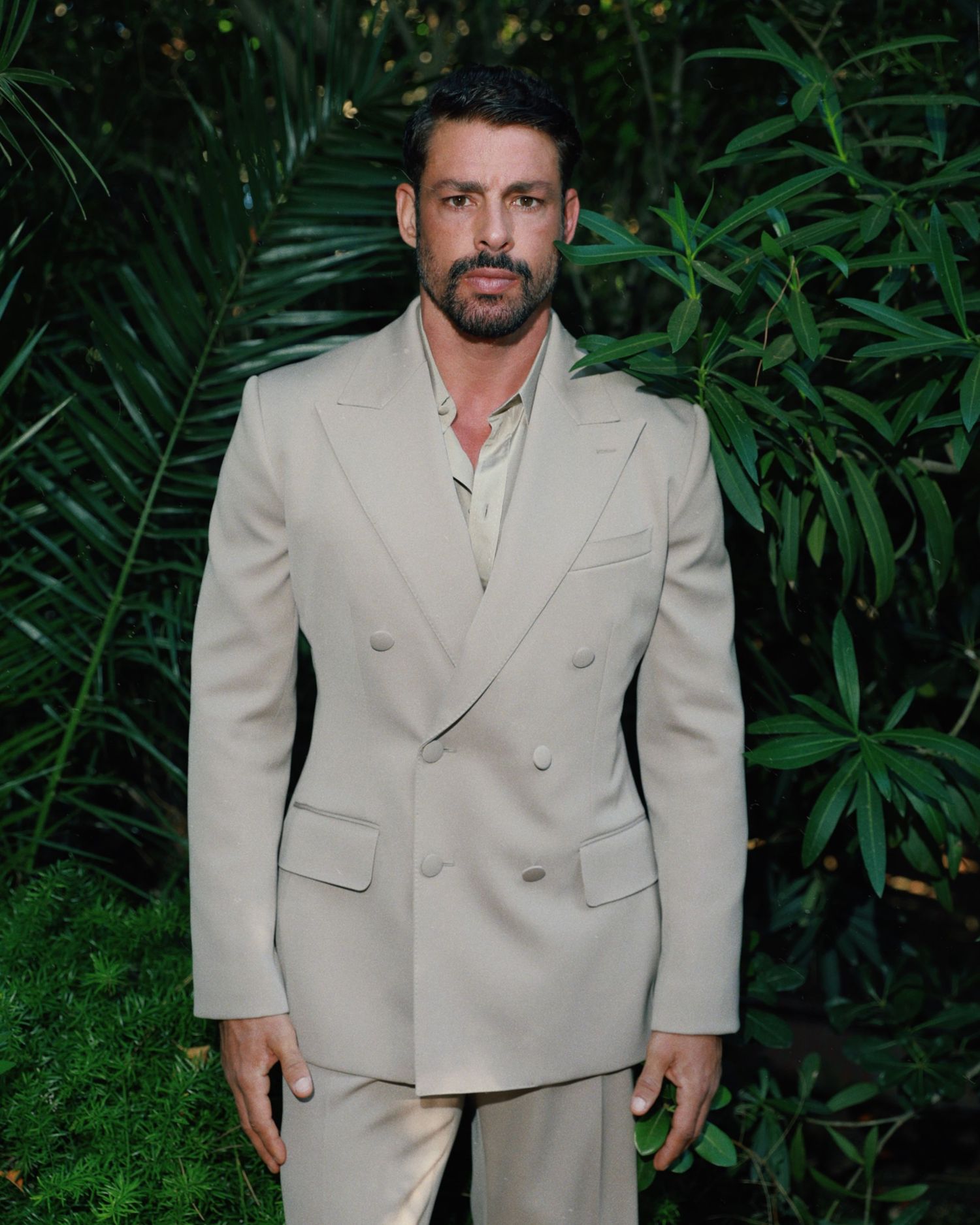 Person wearing a beige double-breasted suit poses against tropical foliage background in outdoor setting.