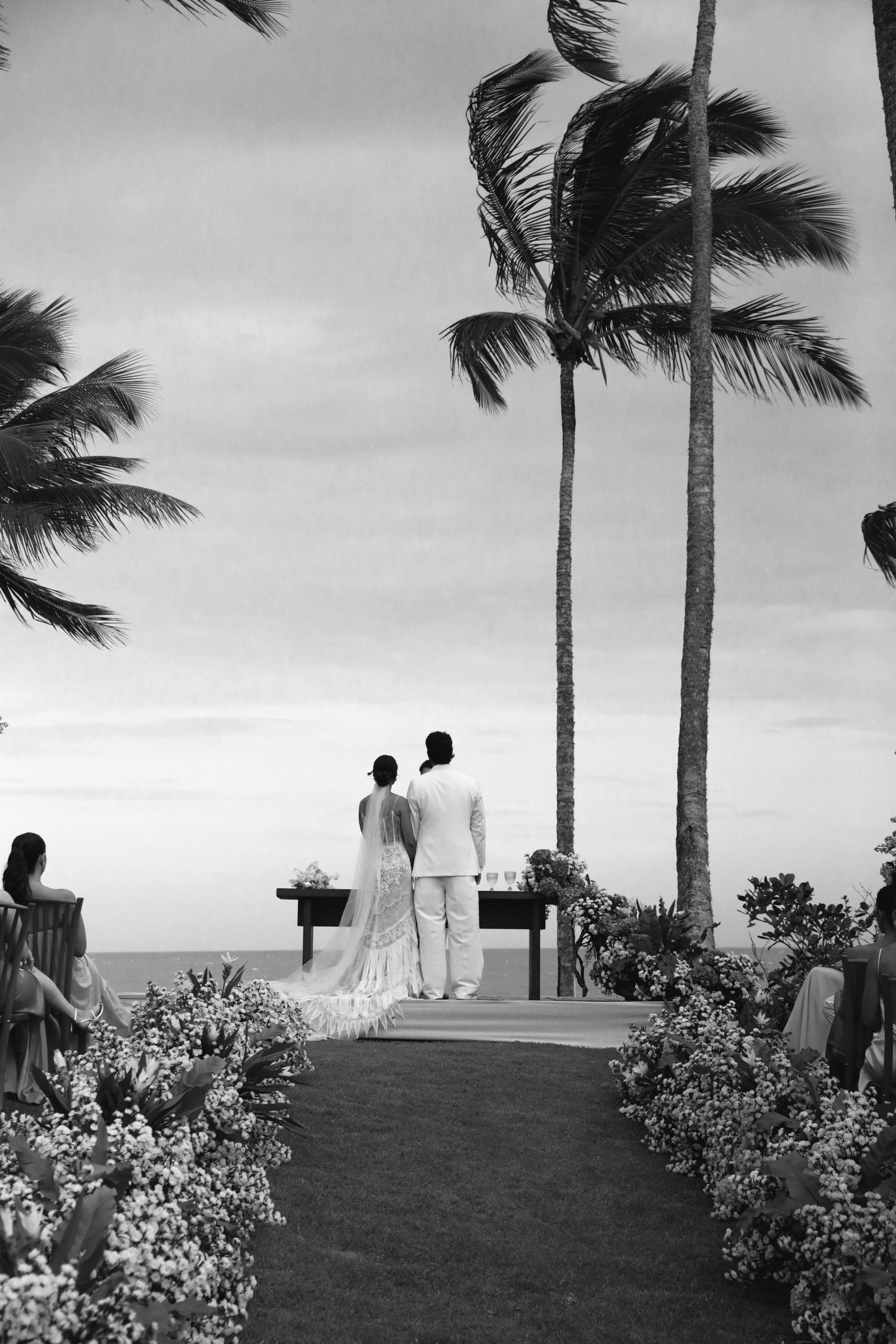 Black and white wedding ceremony overlooking the ocean with palm trees swaying in the wind and flower-lined aisle.