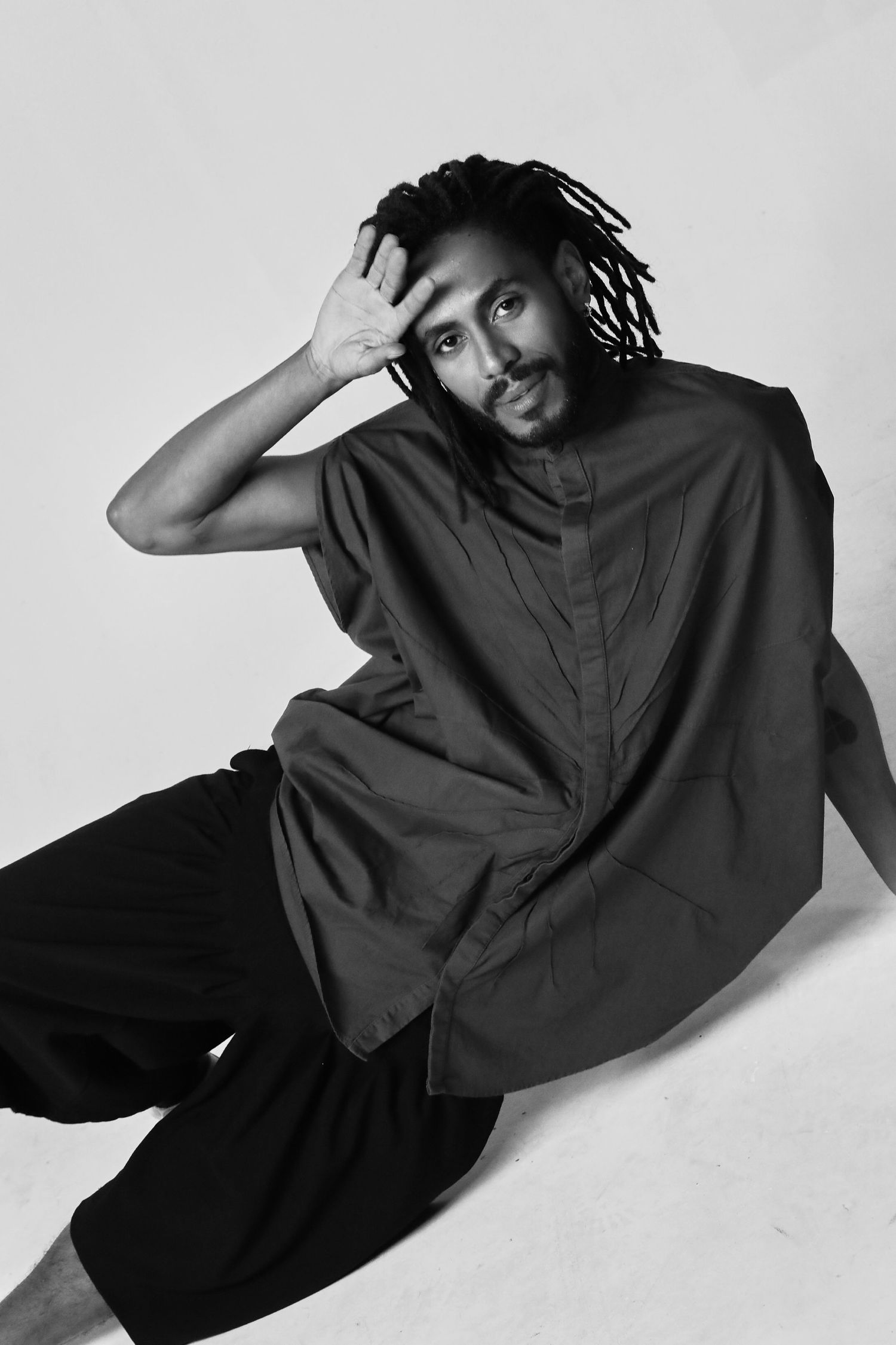 Artistic black and white portrait of a person with dreadlocks in dark clothing making a hand gesture.