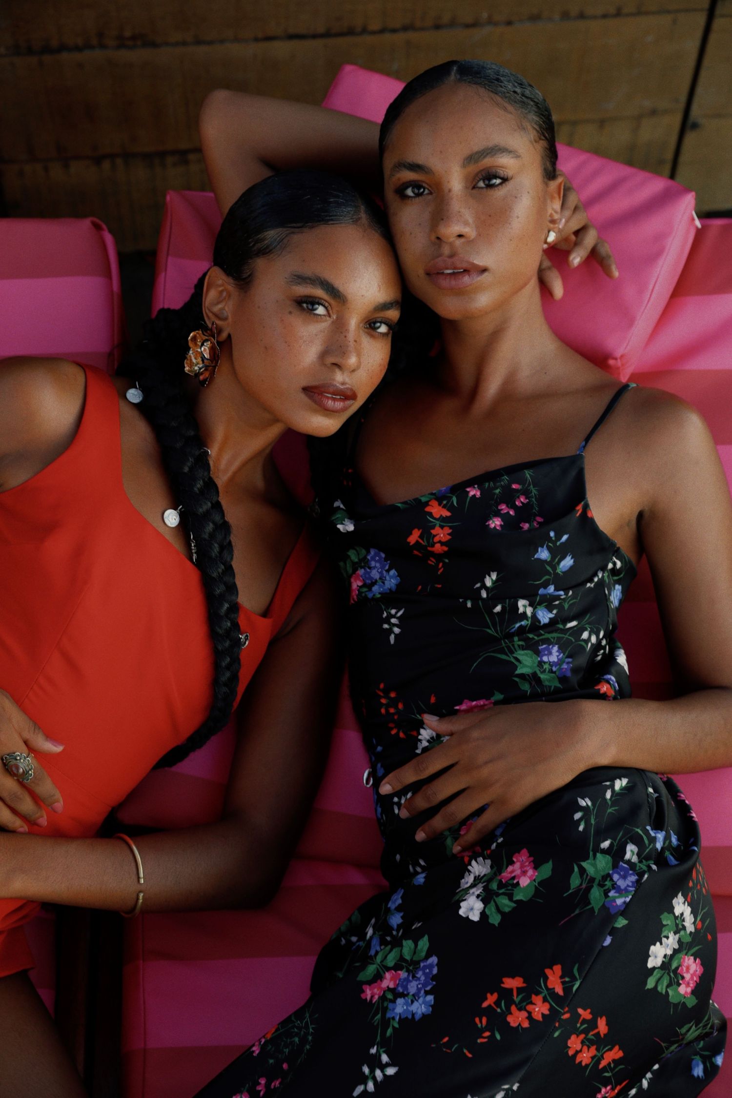 Models in red and floral dresses pose against pink seating and wooden background.