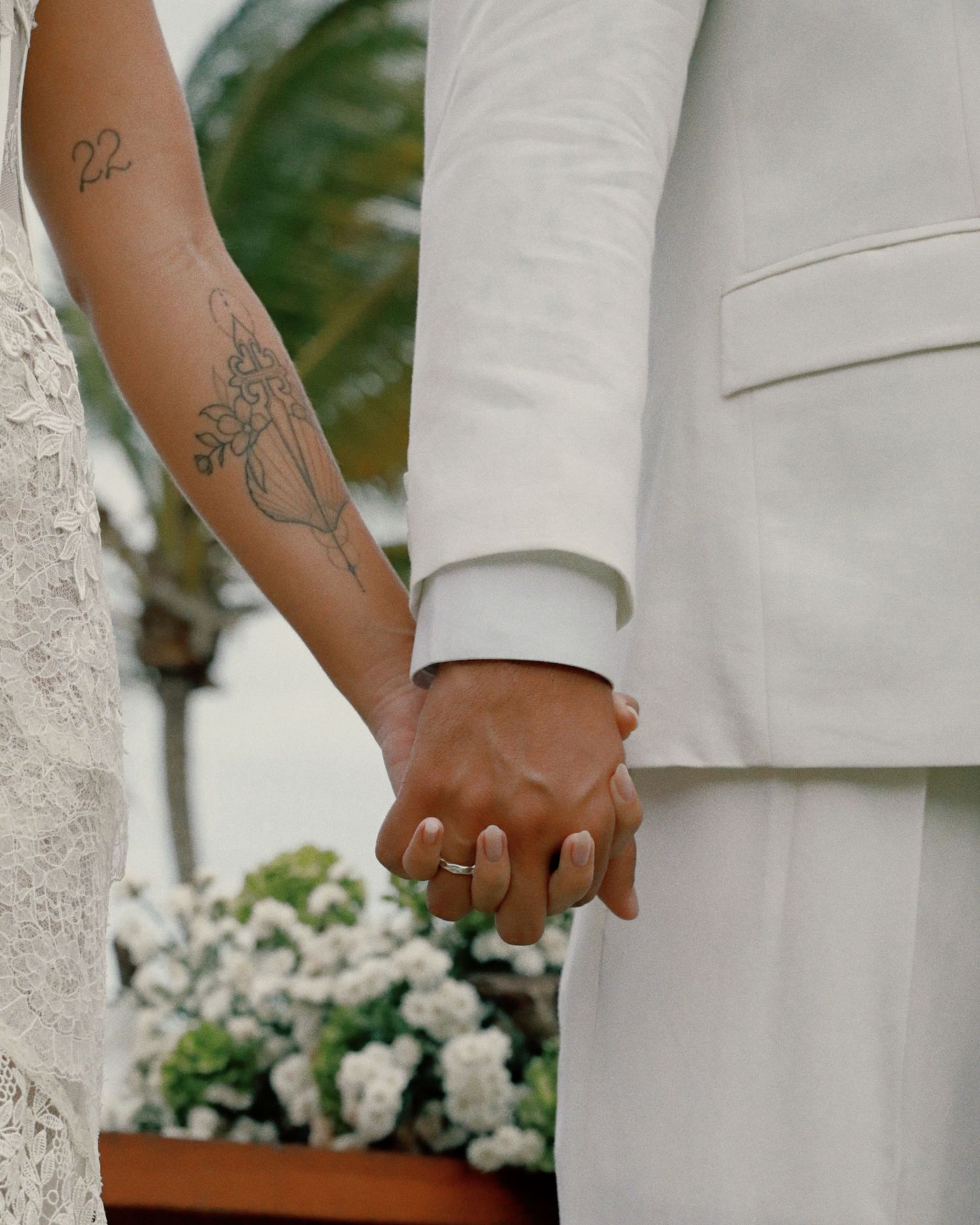 Close-up of newlywed couple holding hands with white flowers and tattoo visible on bride's arm.