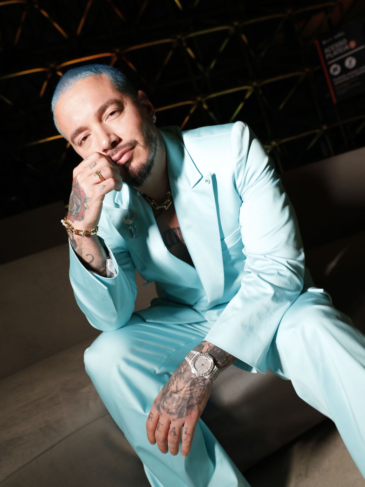 A person wearing a light turquoise suit sits casually in a dimly lit space with tattooed arms resting on their knee.
