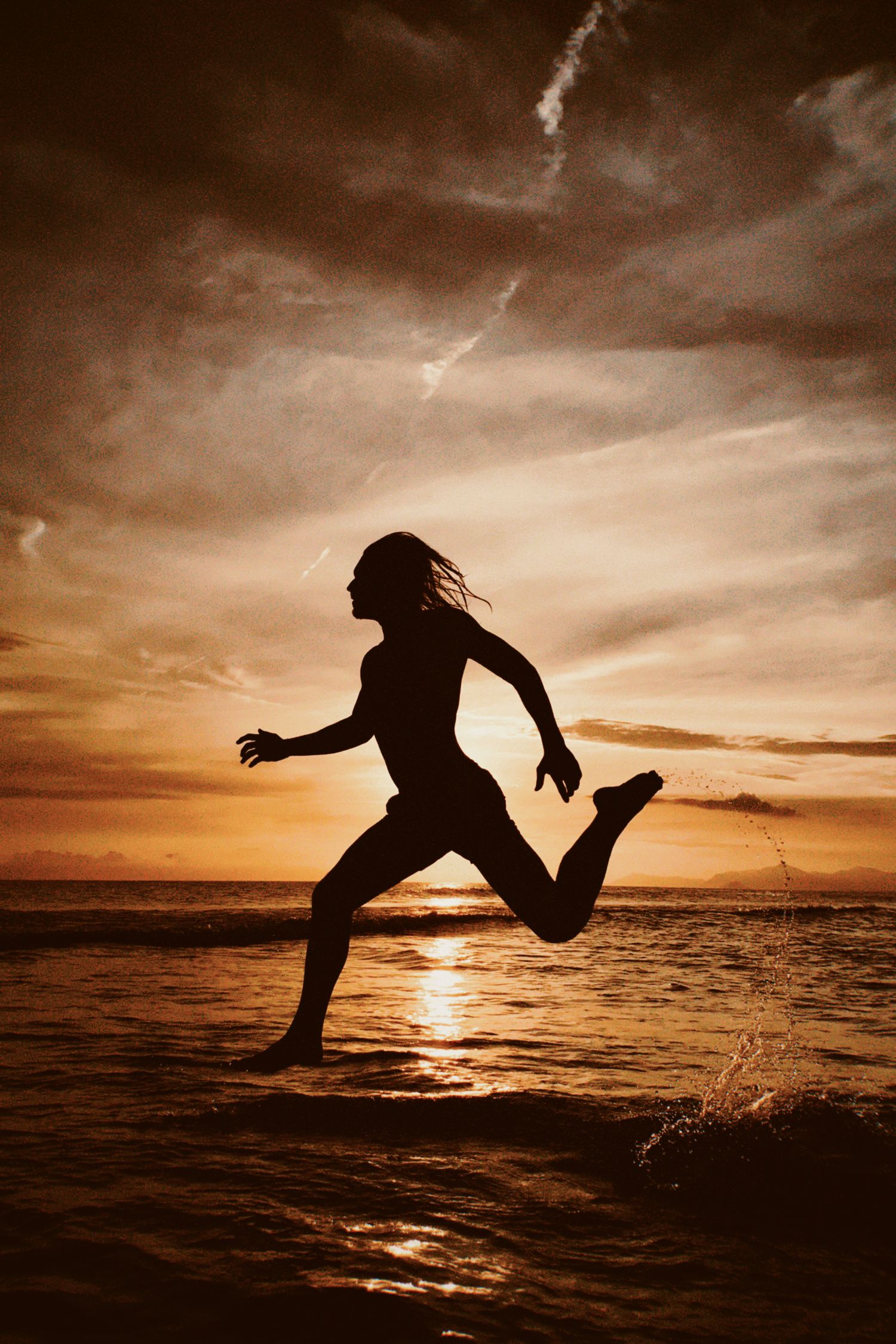 Silhouette of person running through shallow ocean water at sunset with dramatic orange sky and clouds.