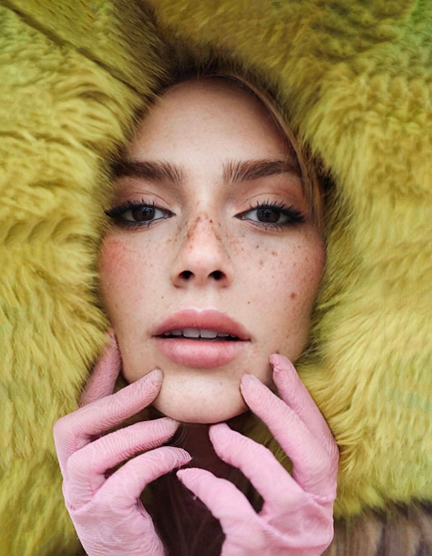 Close up portrait against yellow furry background with pink gloves touching face and natural freckled makeup look.