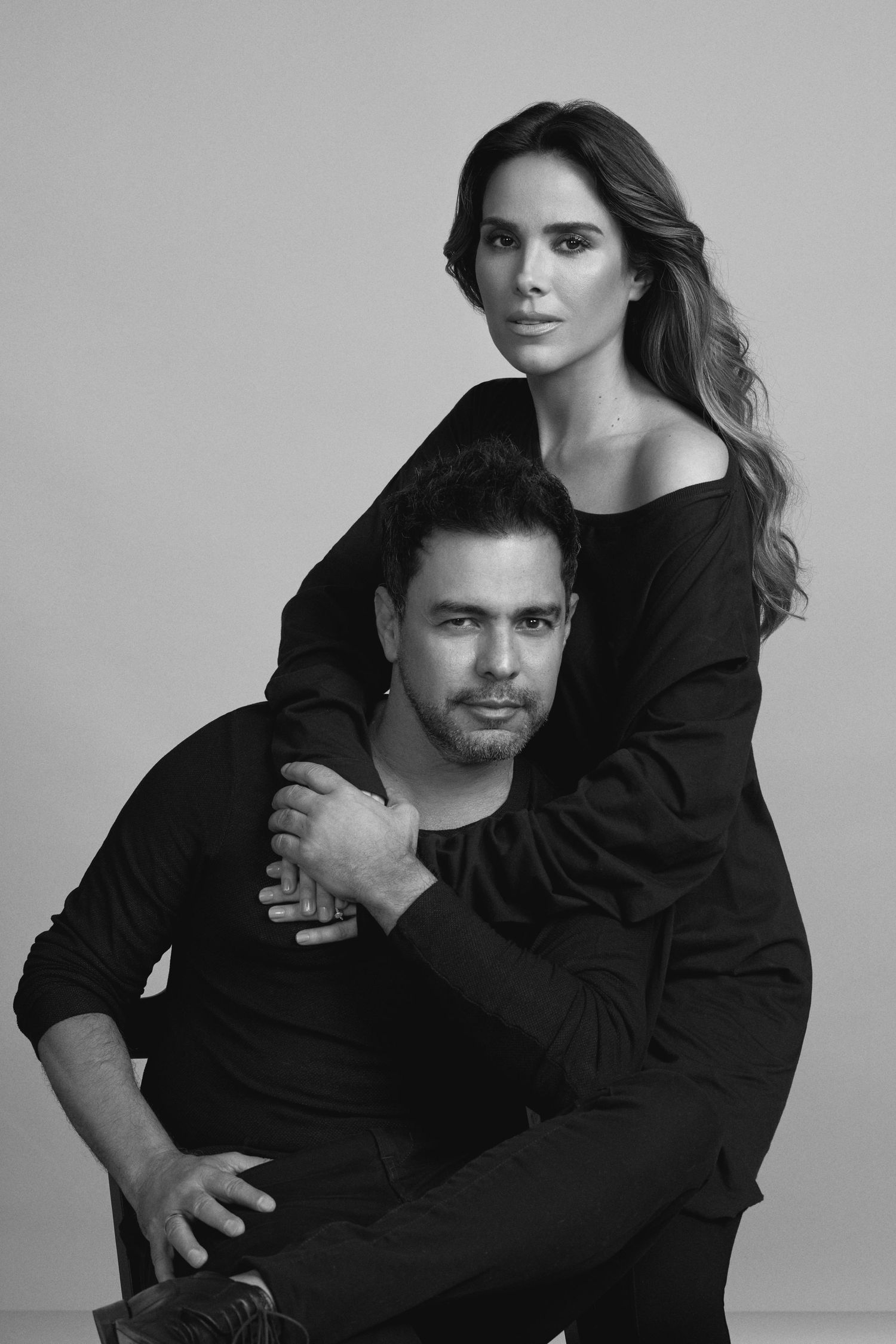 A black and white studio portrait of a couple in dark clothing embracing in an artistic pose.