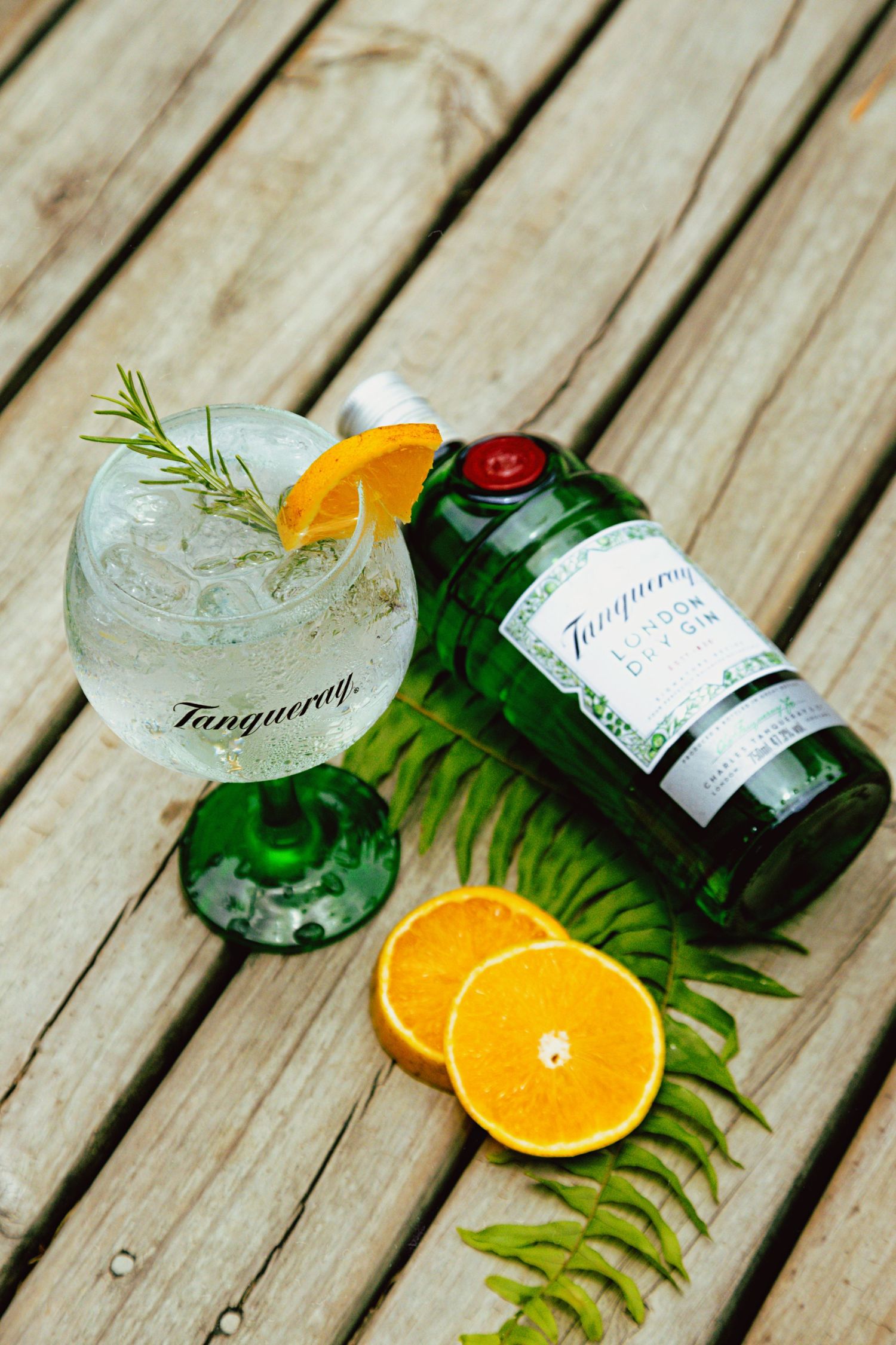 Tanqueray gin cocktail with orange slices and rosemary garnish on wooden deck with tropical leaves.