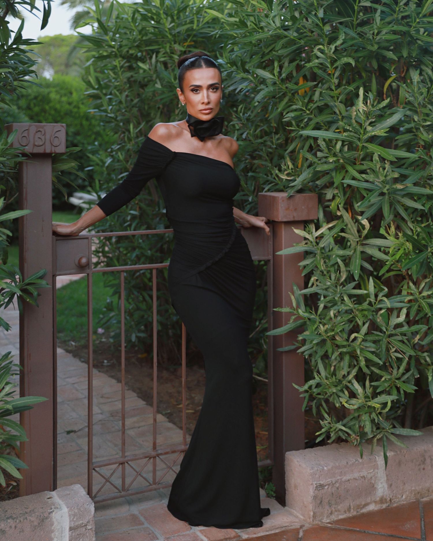 Model in form-fitting black gown poses elegantly by rustic garden gate.