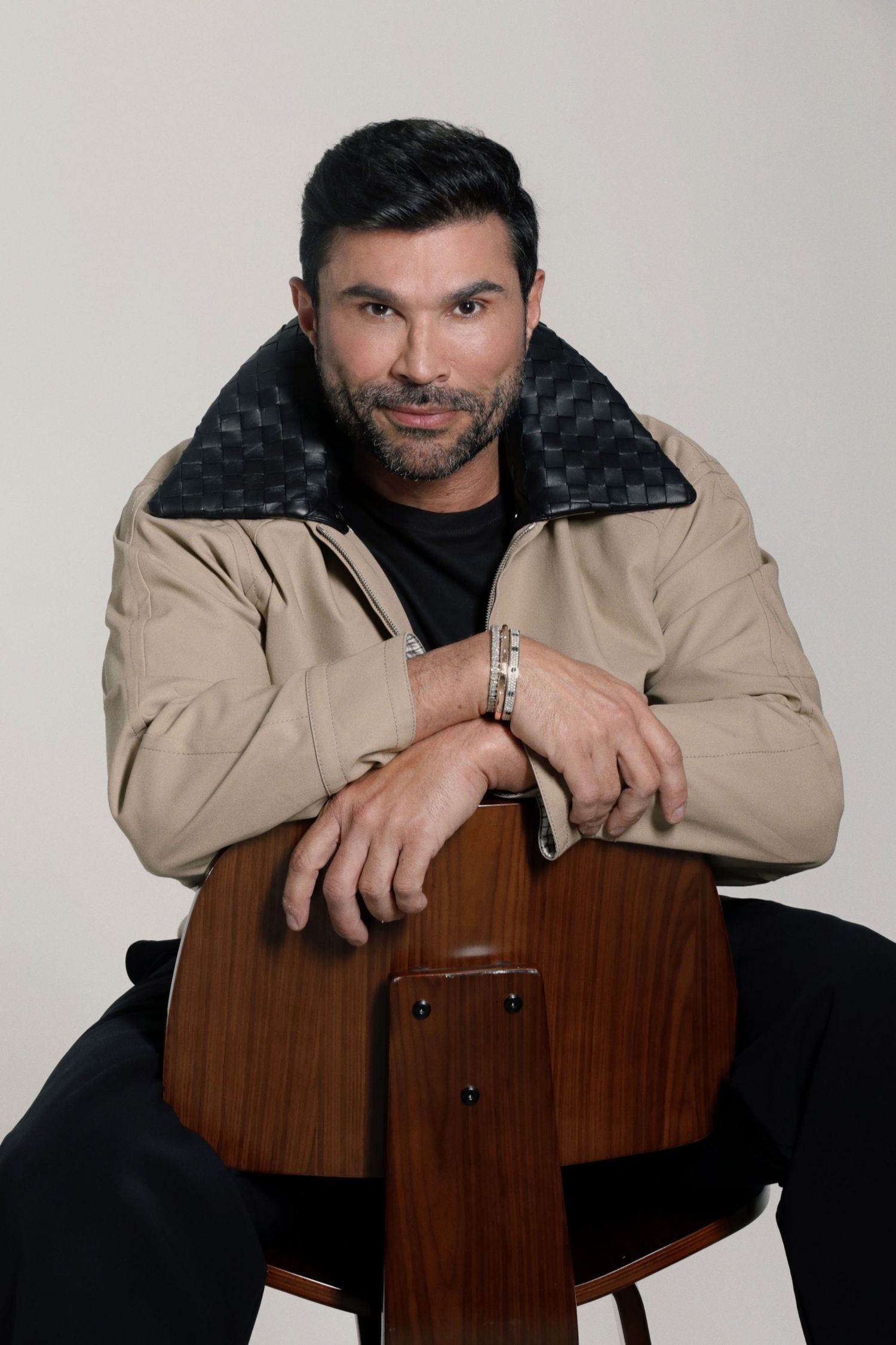Man in beige jacket poses confidently on wooden chair against neutral background.
