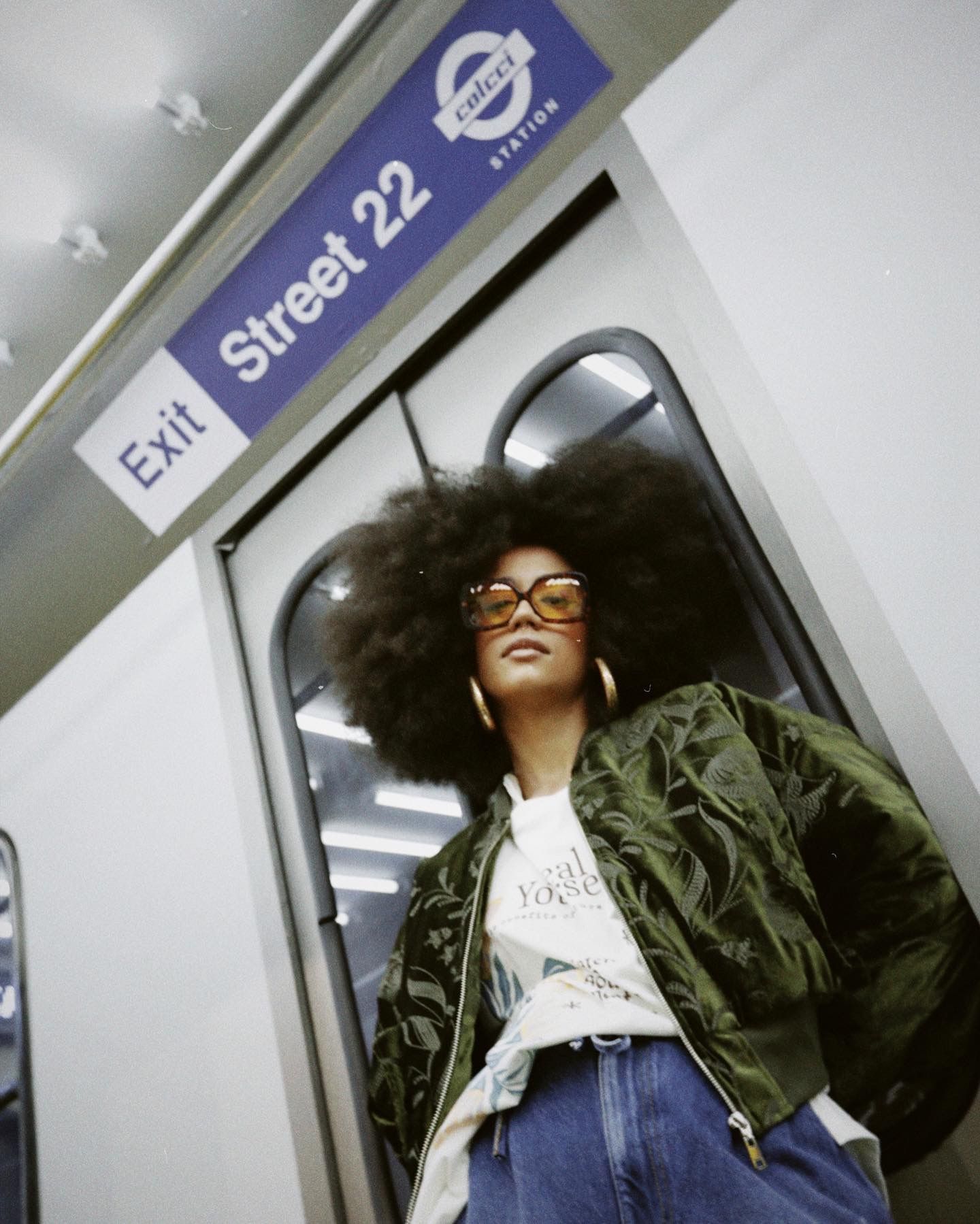 A stylish commuter wearing glasses and natural hair poses against the subway door near a Street 22 exit sign.