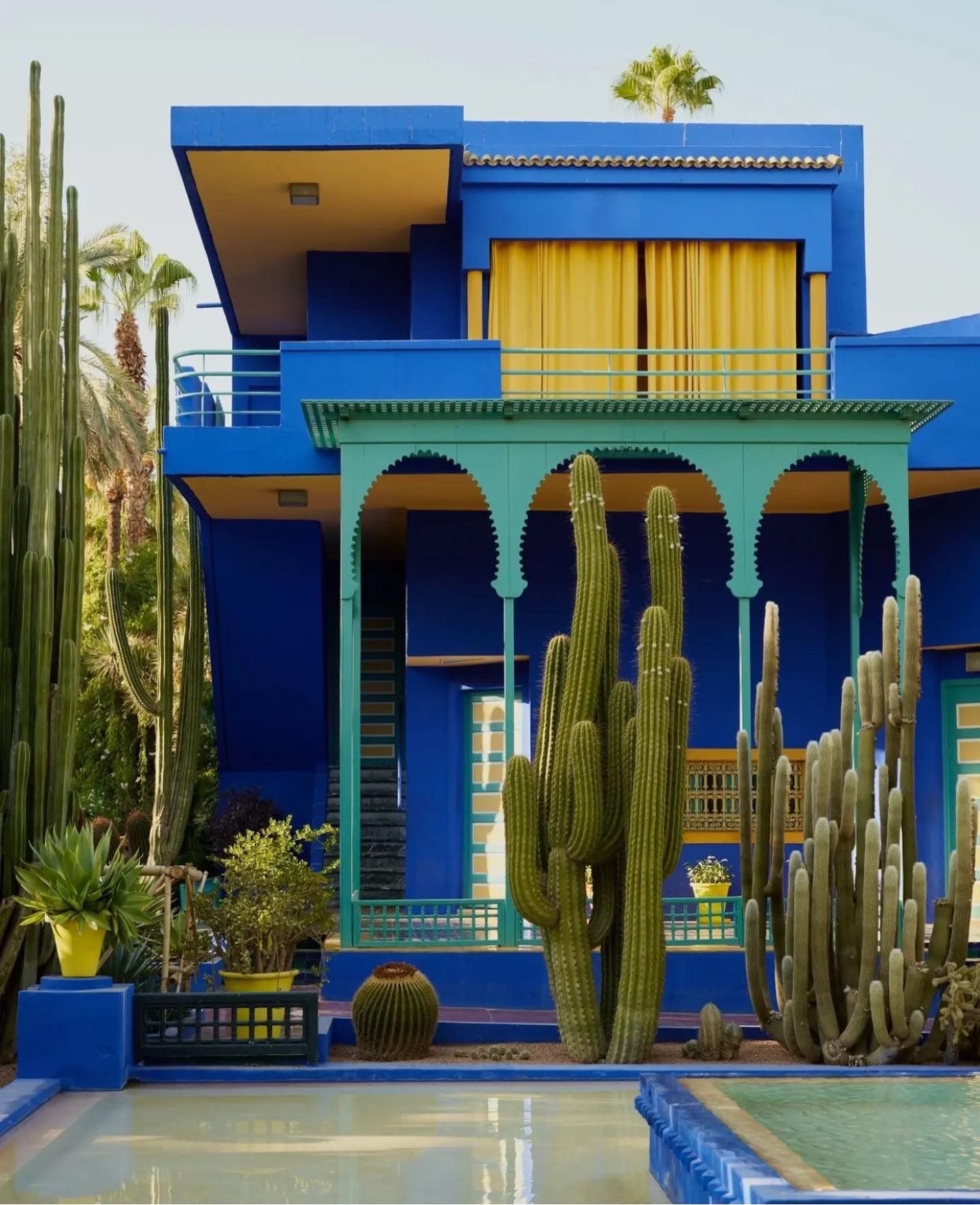 Vibrant blue and yellow Moroccan-style house with green arches and tall cactus plants surrounding a pool in a desert setting.
