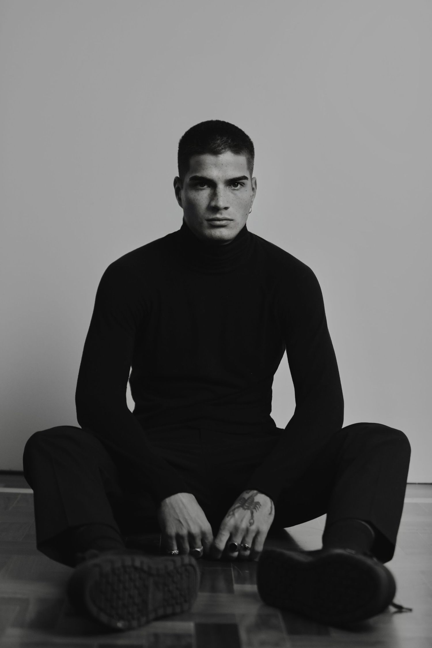 Black and white portrait of a person in a dark turtleneck sweater sitting cross-legged on the floor.