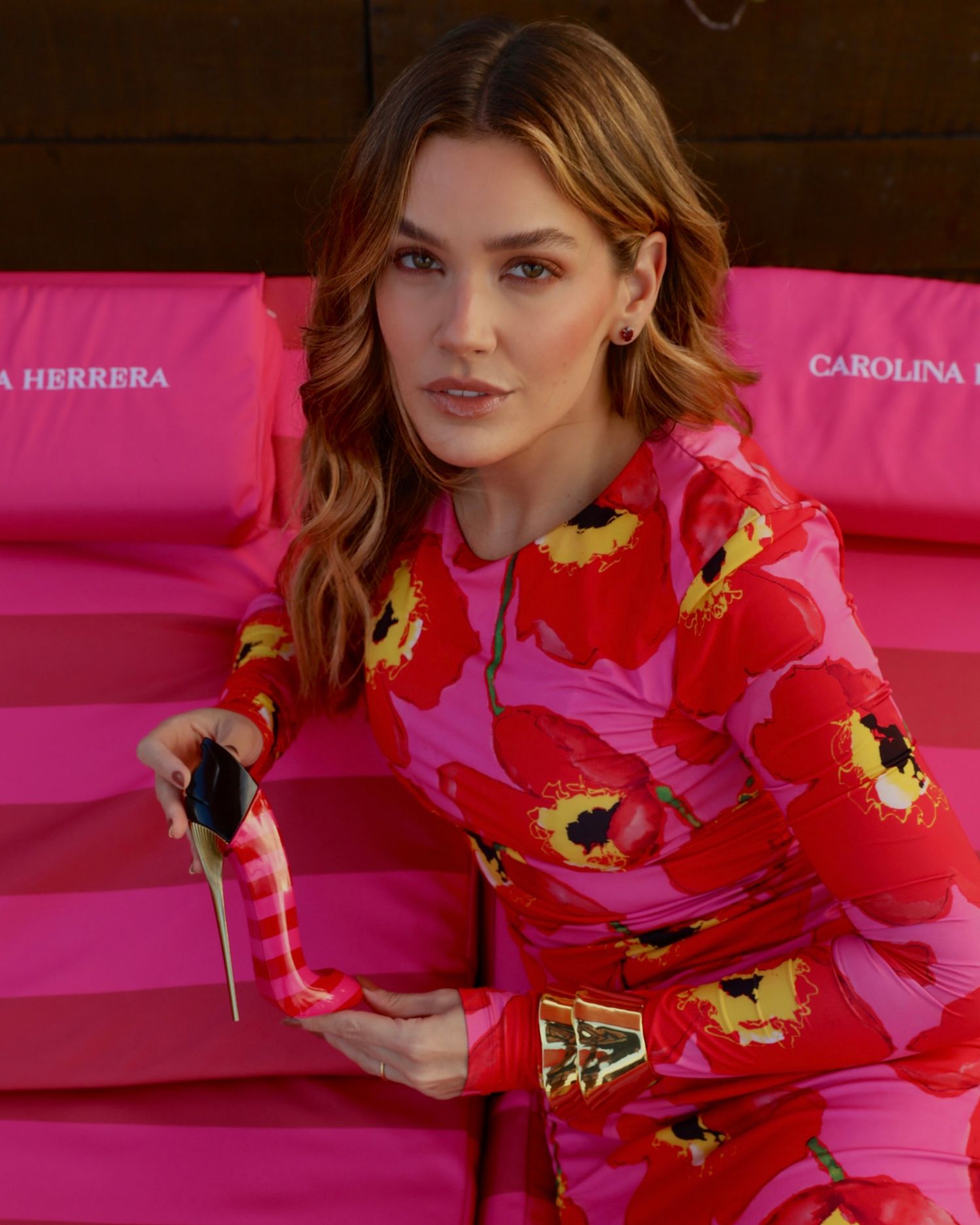 Fashion model poses in vibrant red and pink floral print dress against pink Carolina Herrera branded chairs.