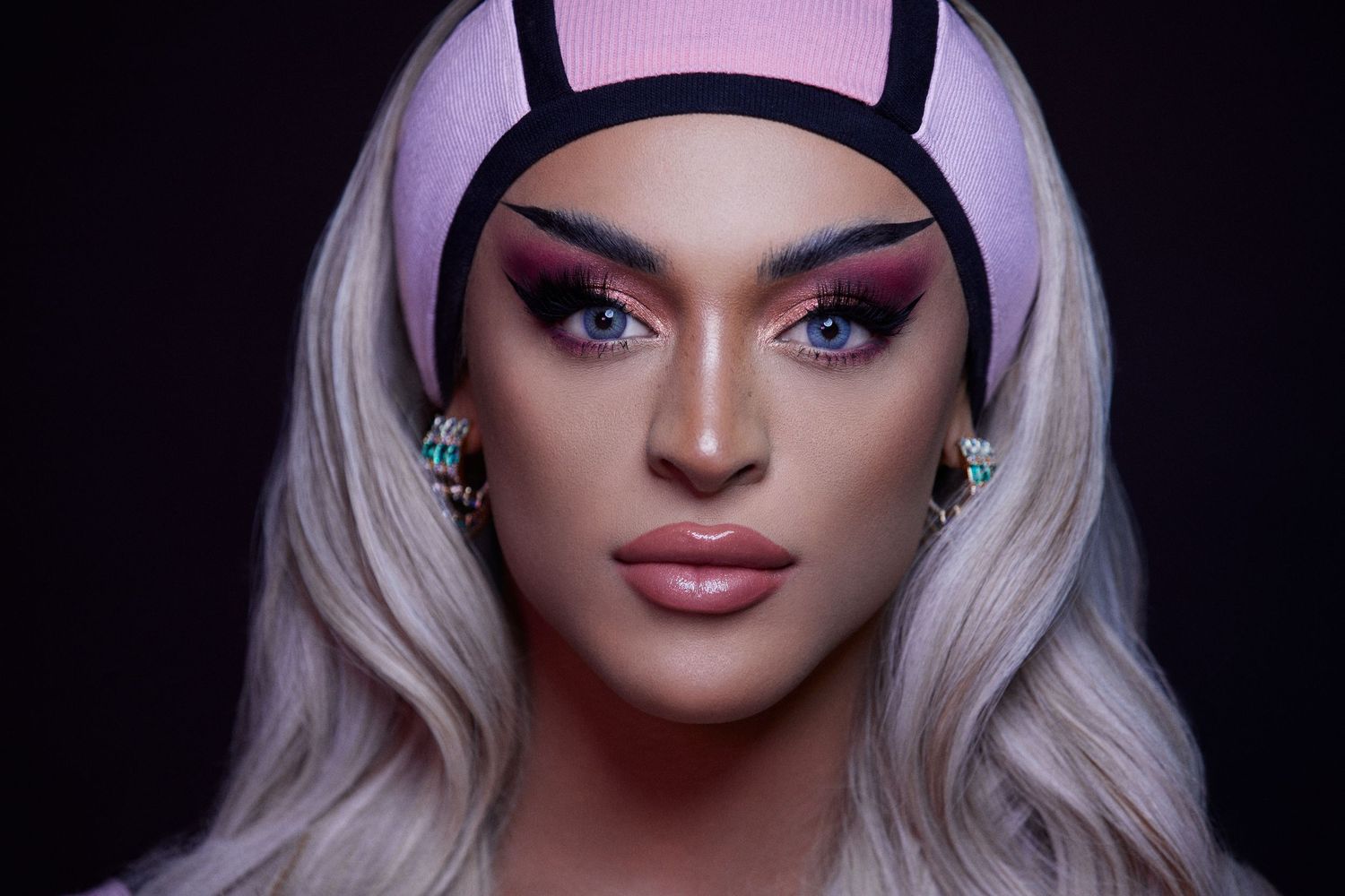 Glamorous portrait of a drag performer with platinum blonde hair wearing black horns and dramatic pink eye makeup in low lighting.