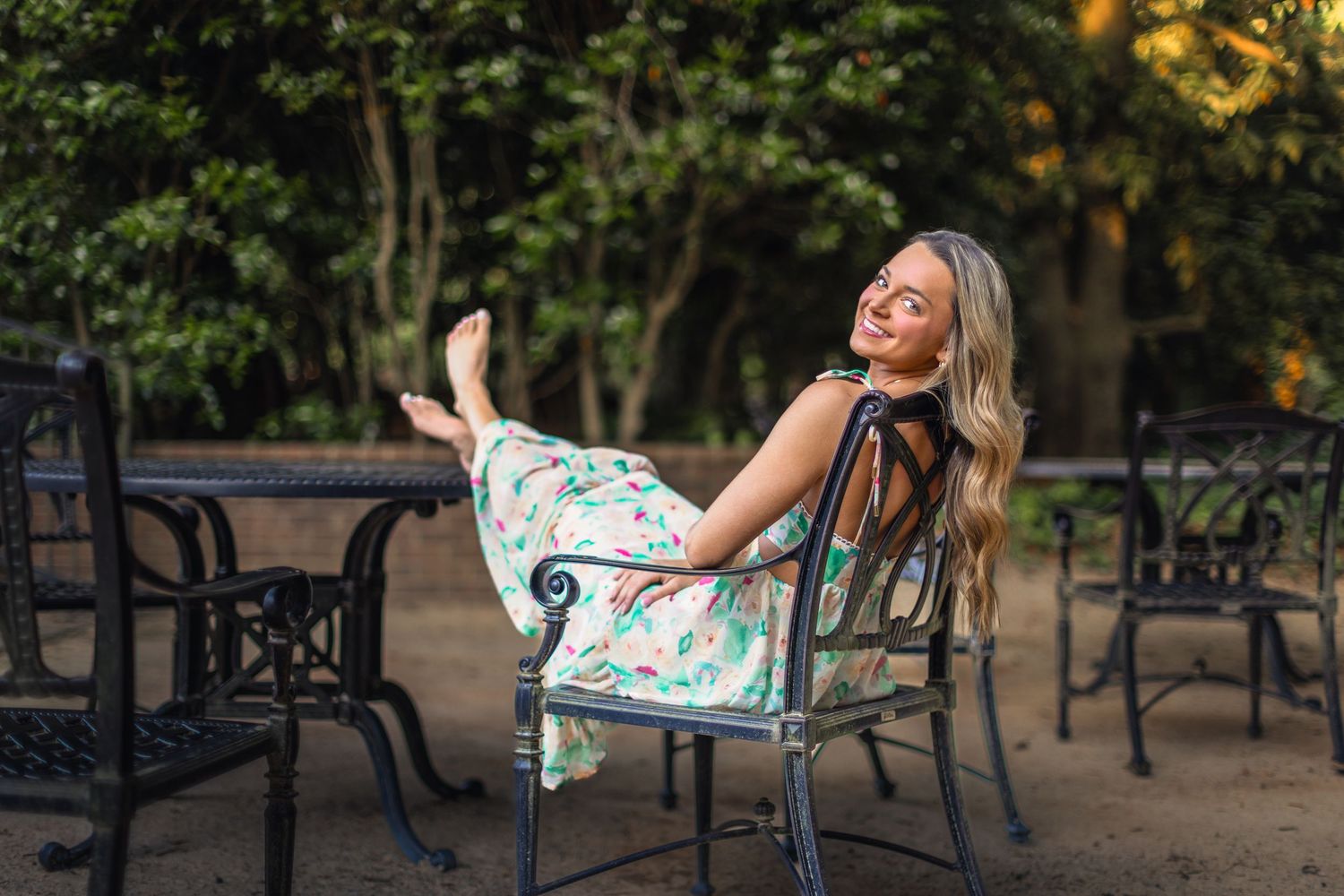A blonde girl in a mulitcolor dress with feet on a table looking back over her shoulder while smiling