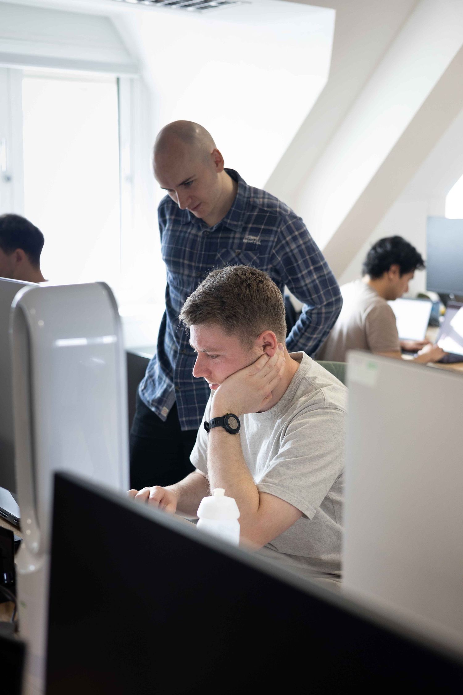 man working at desk looking at computer screen while a man dressed in a shirt sits over him