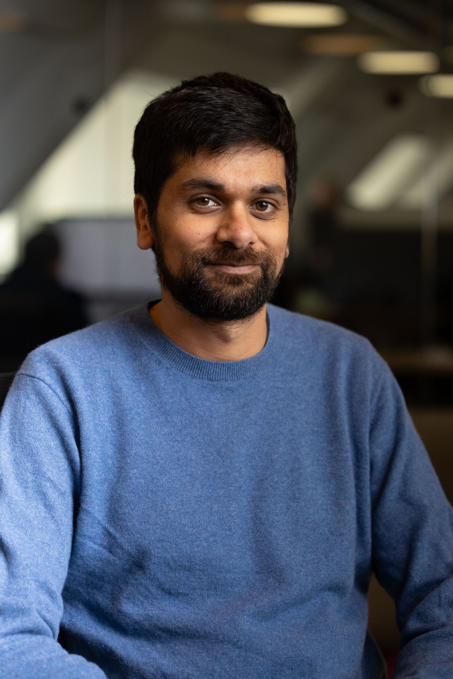 A head shot of a Indian man in blue shirt with the office behind him