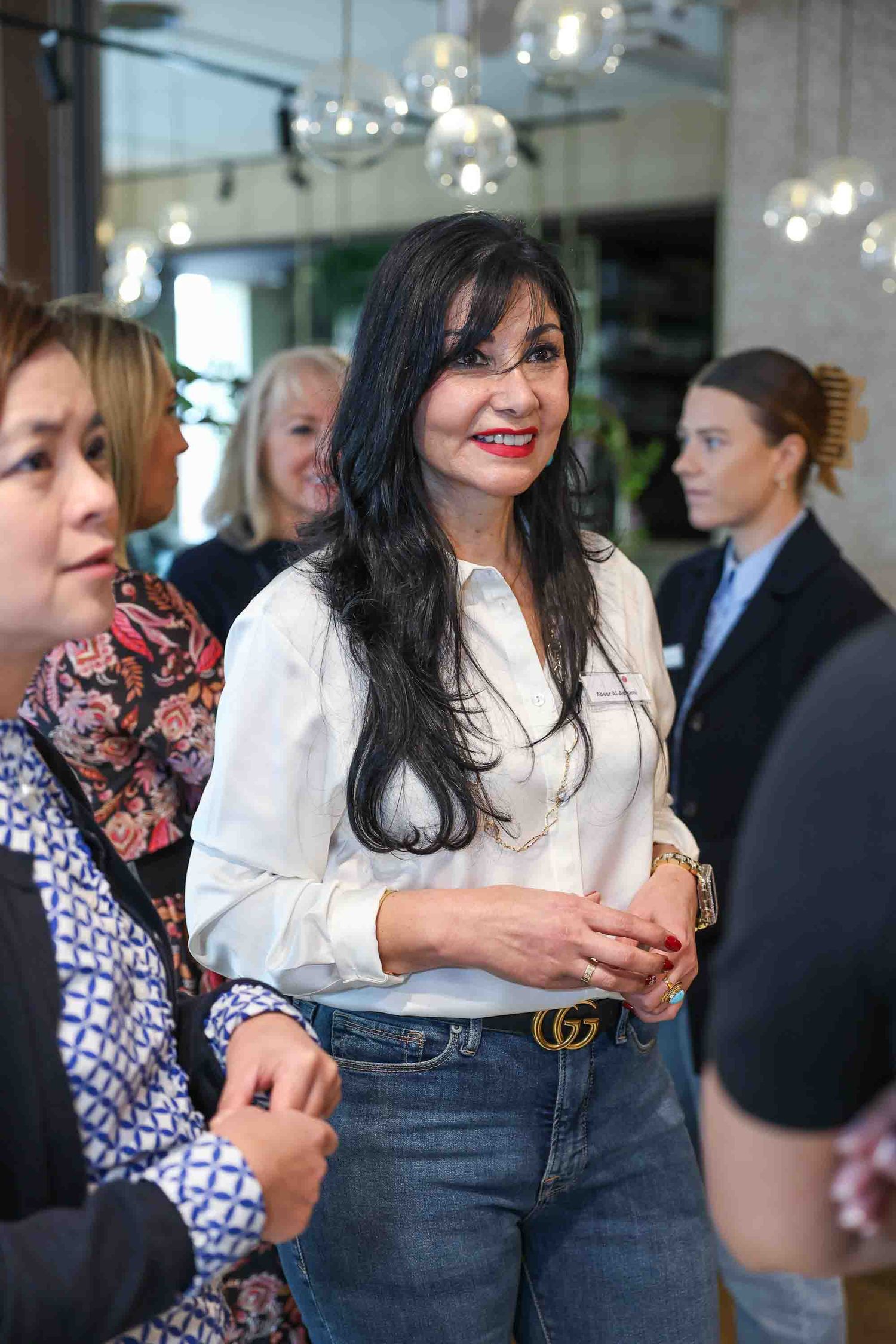 A woman in a white top and blue jeans smiling in a crowded business network meeting
