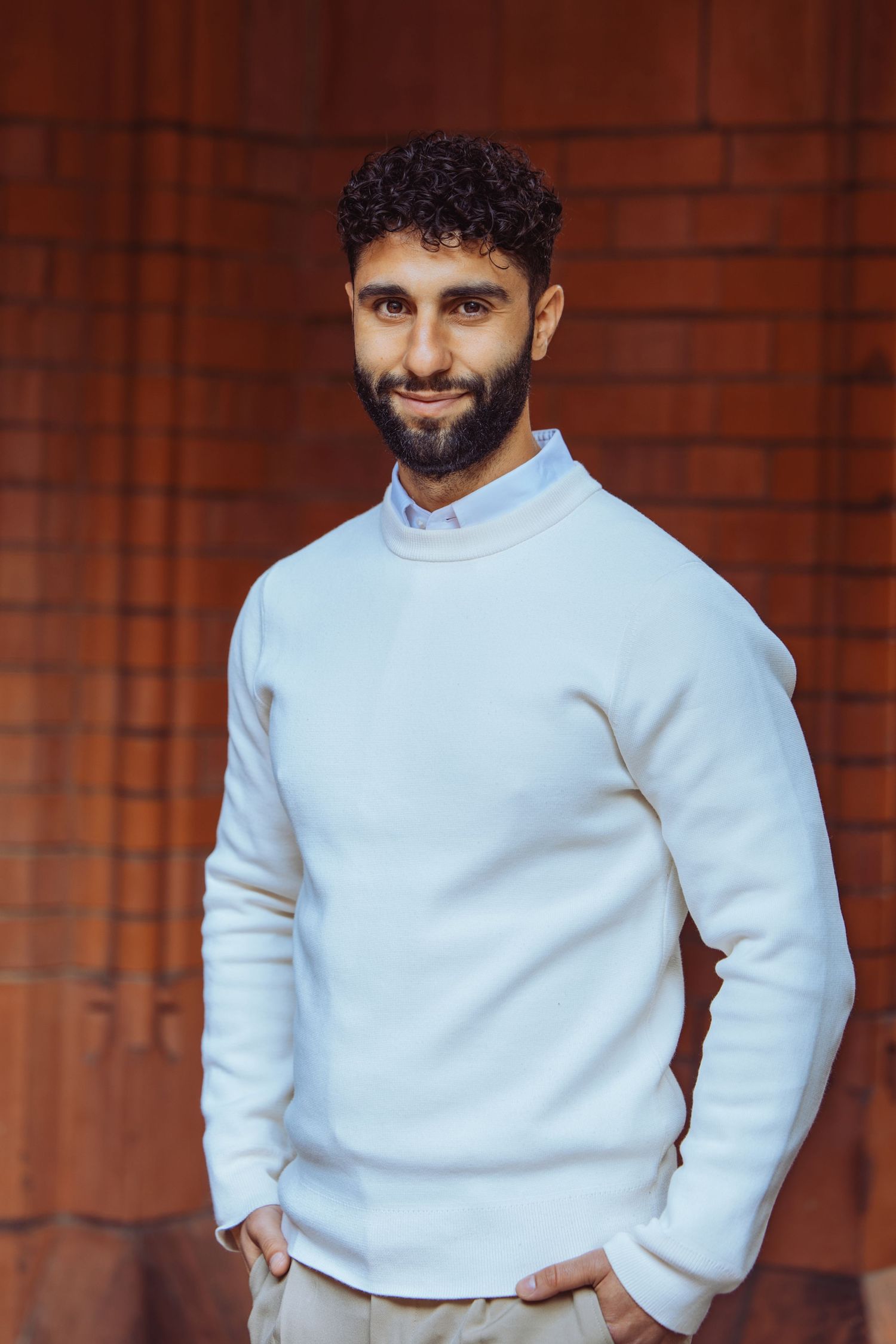 A Indian man with a white jumper against a orange brick wall